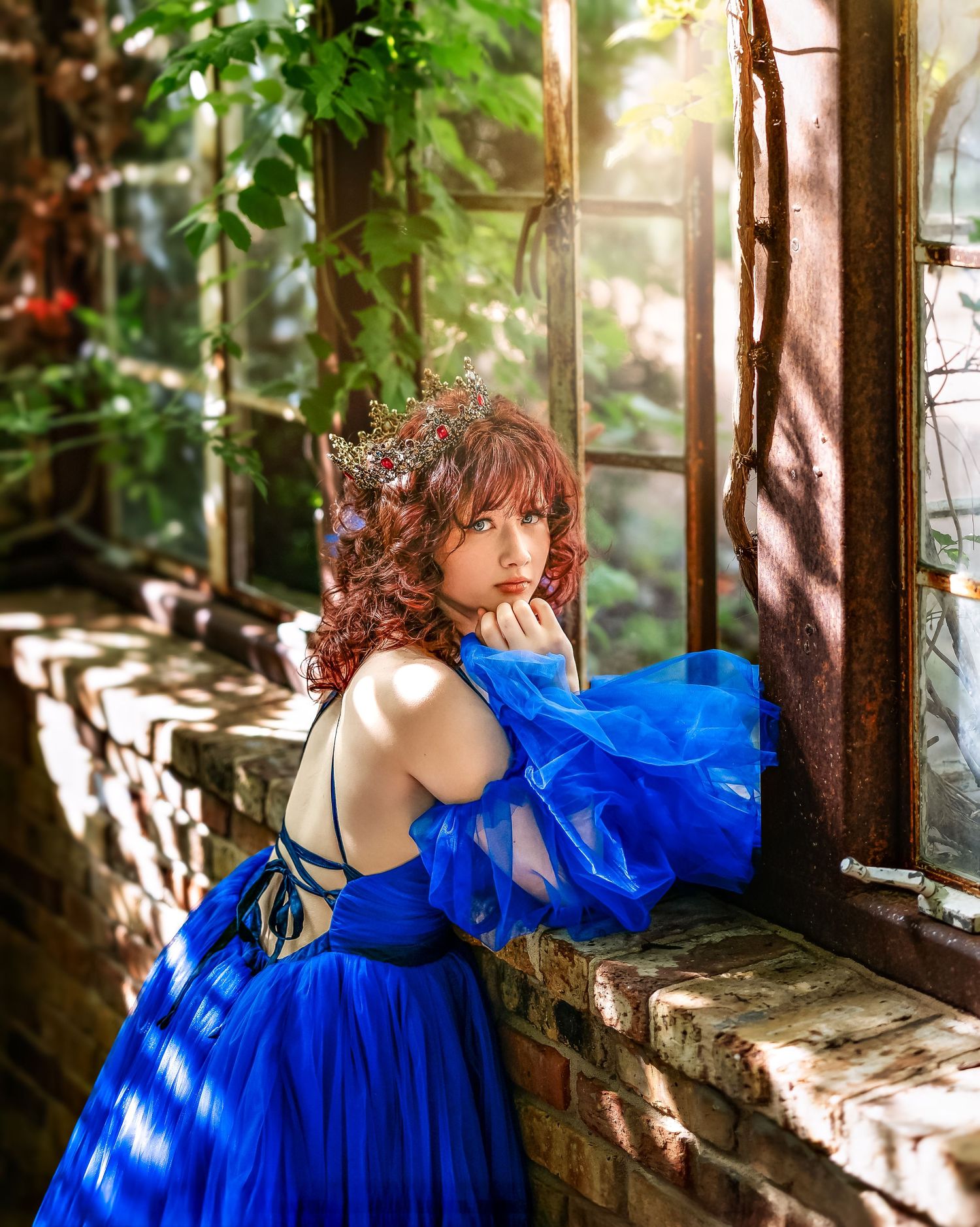 Young woman in a royal blue ball gown with floral crown poses by a sunlit greenhouse window.