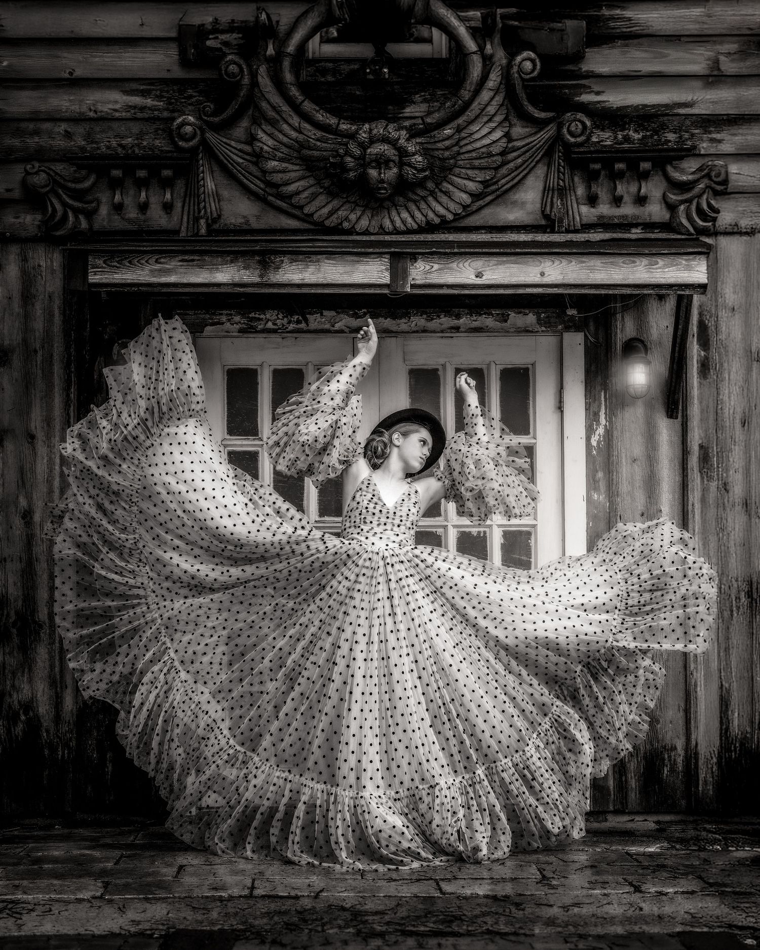 Black and white photo of a woman in a flowing polka dot dress dancing gracefully before an ornate wooden building.