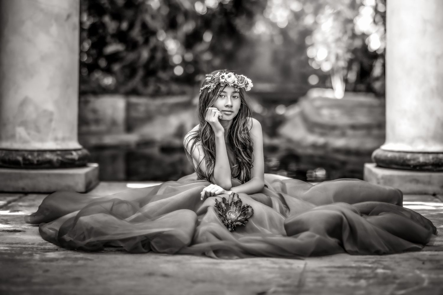 Black and white photo of a young woman in a floral crown and flowing gown seated gracefully outdoors.
