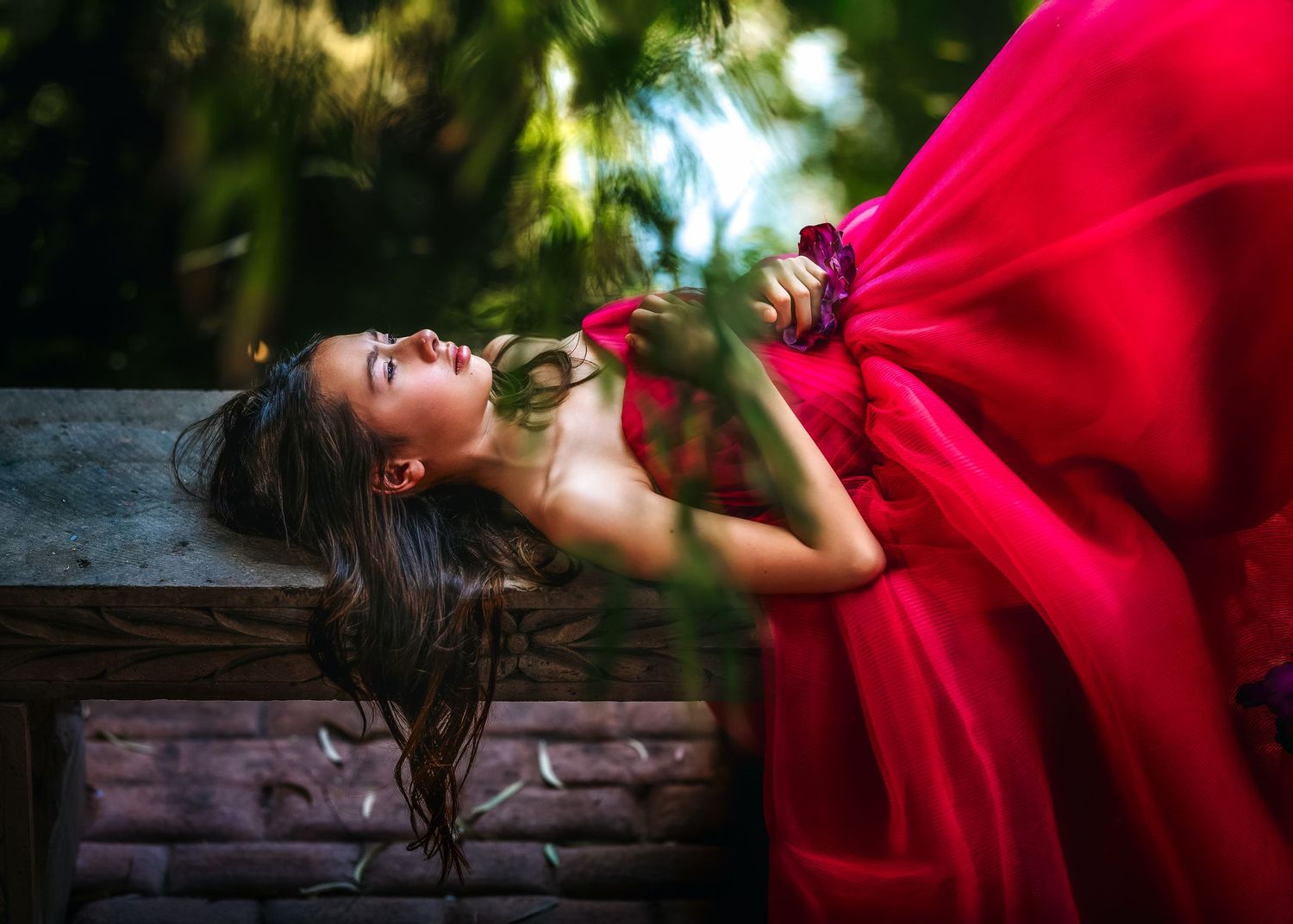 A young woman in a flowing red gown reclines gracefully on a wooden bench surrounded by lush green foliage.
