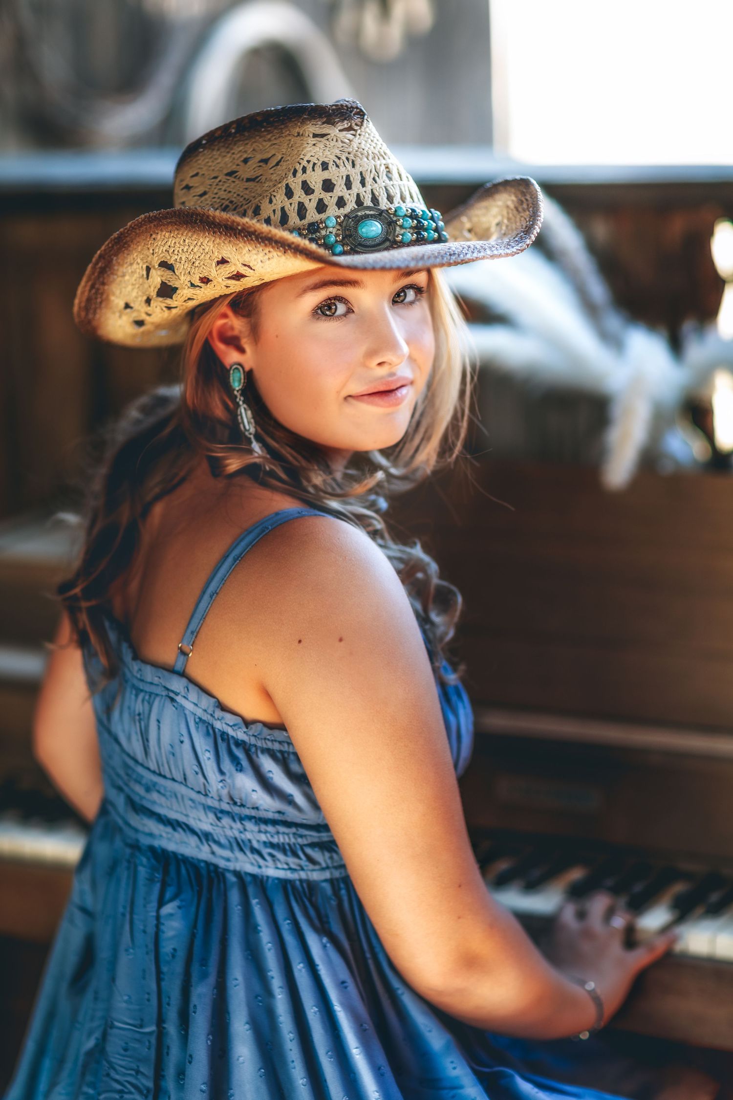 Young woman in blue dress and turquoise cowboy hat sitting at vintage piano.