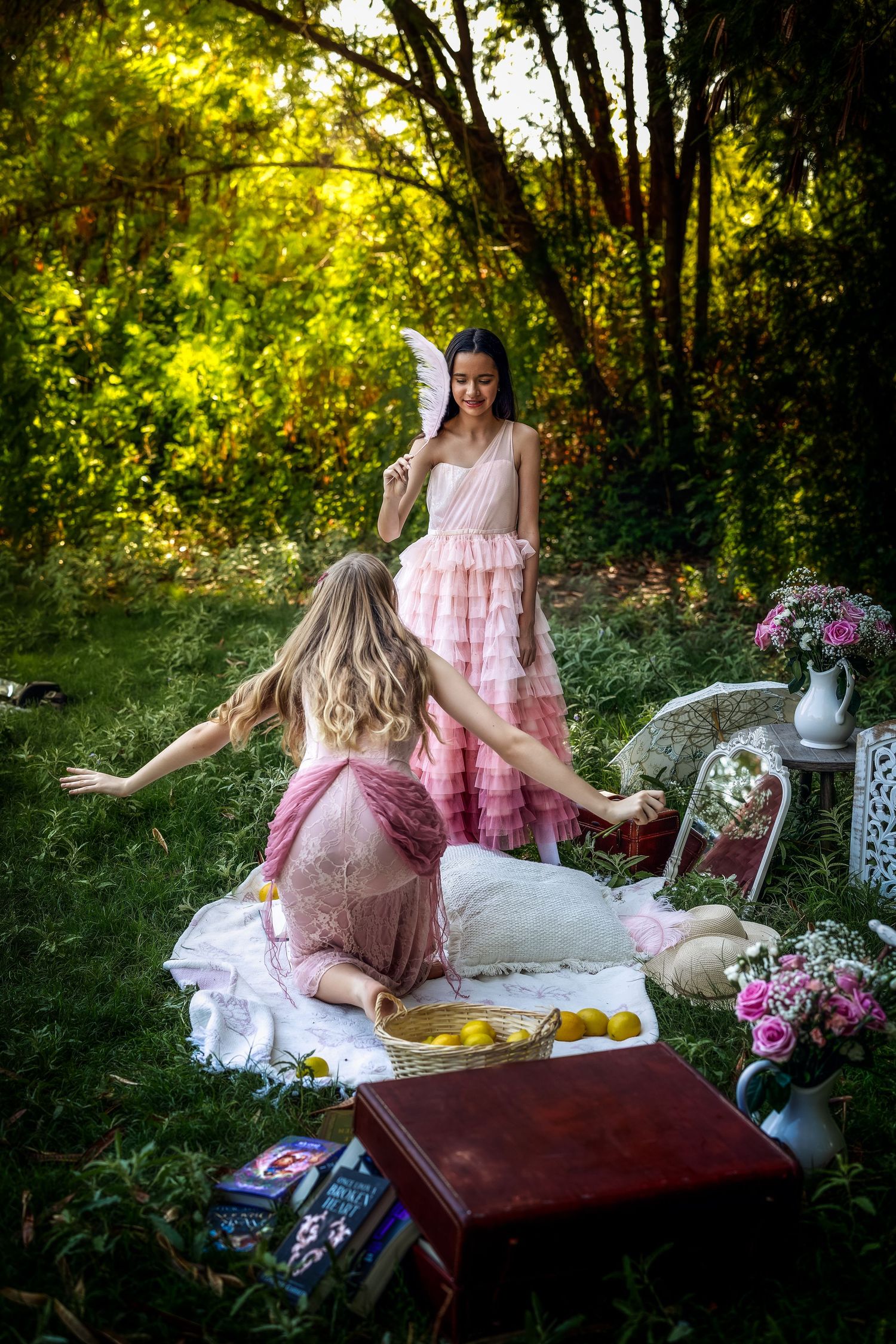 Two women in pink dresses enjoying a garden picnic surrounded by flowers and books in sunlight.