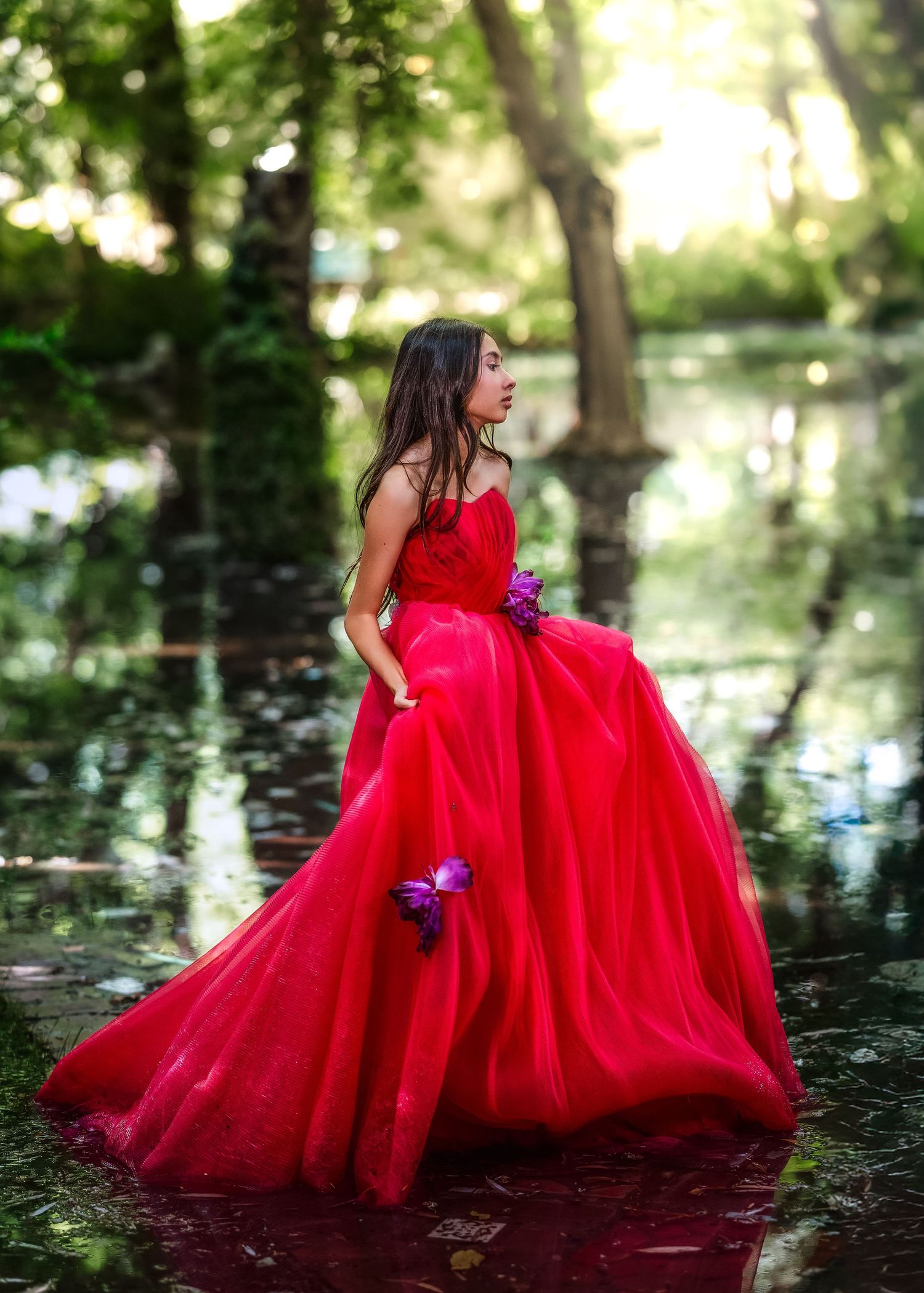Young girl in red tulle ball gown with purple butterflies stands in flooded forest clearing.