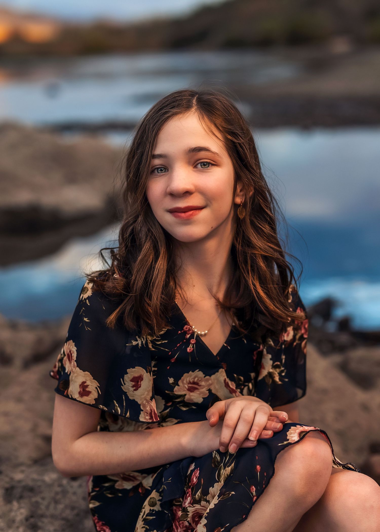 Portrait of a teenage girl in a floral dress sitting on rocks by a reflective blue pond.
