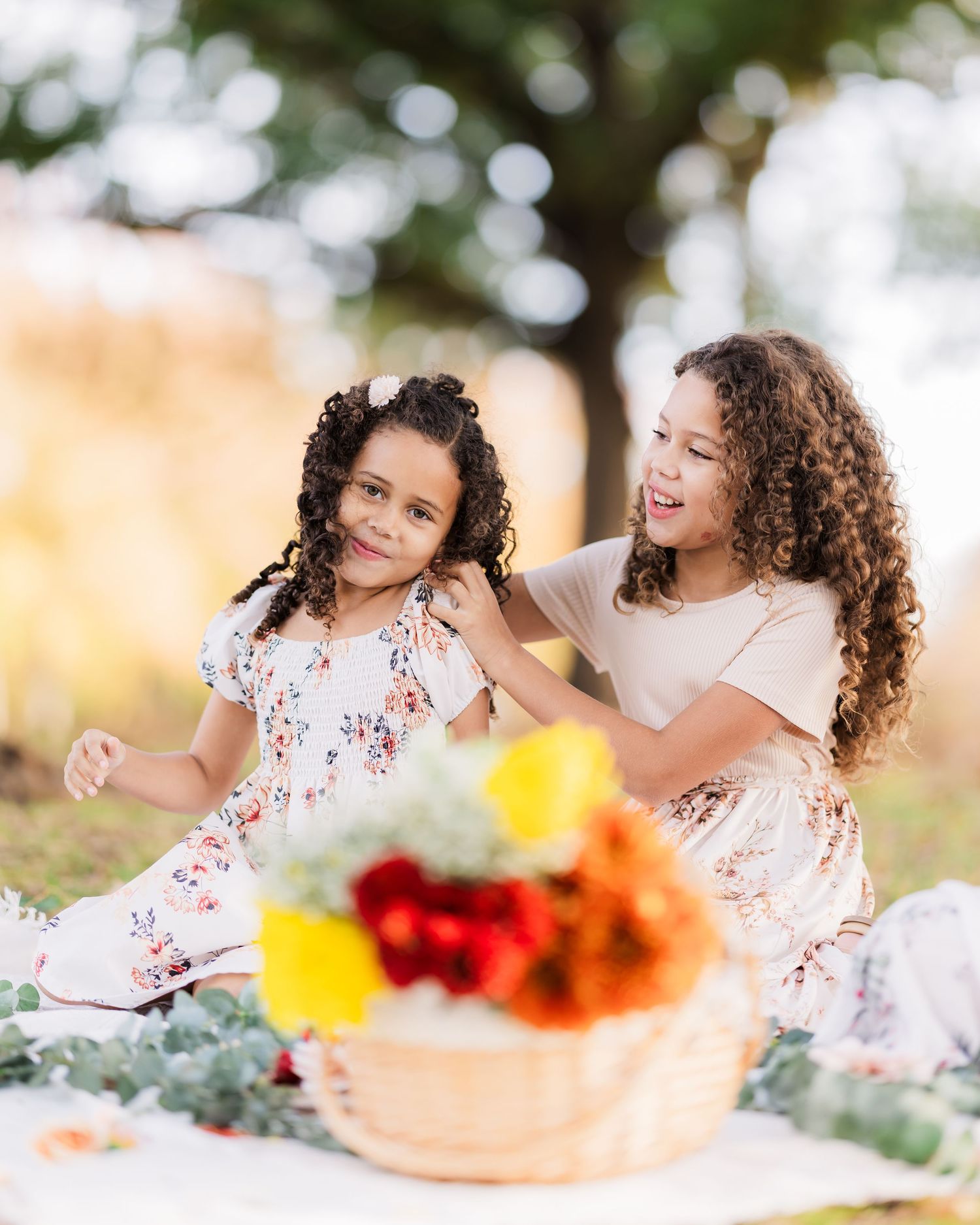 two sisters with beautiful curls sit on blanket in a park with a picnic basket full of flowers in the foreground.