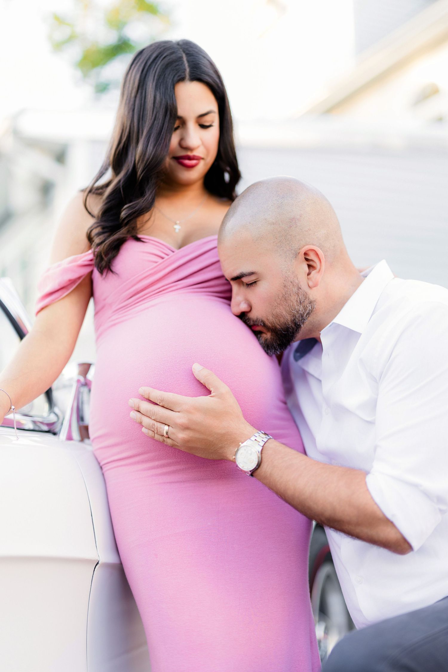 man kissing pregnant female's belly while leaning against white car