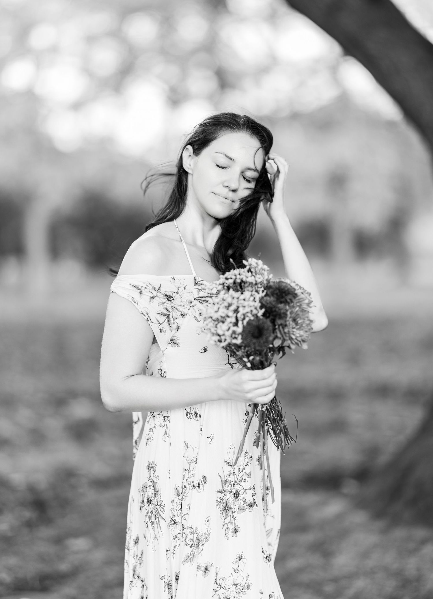 black and white photo of female with her eyes closed holding a bouquet of flowers underneath a tree