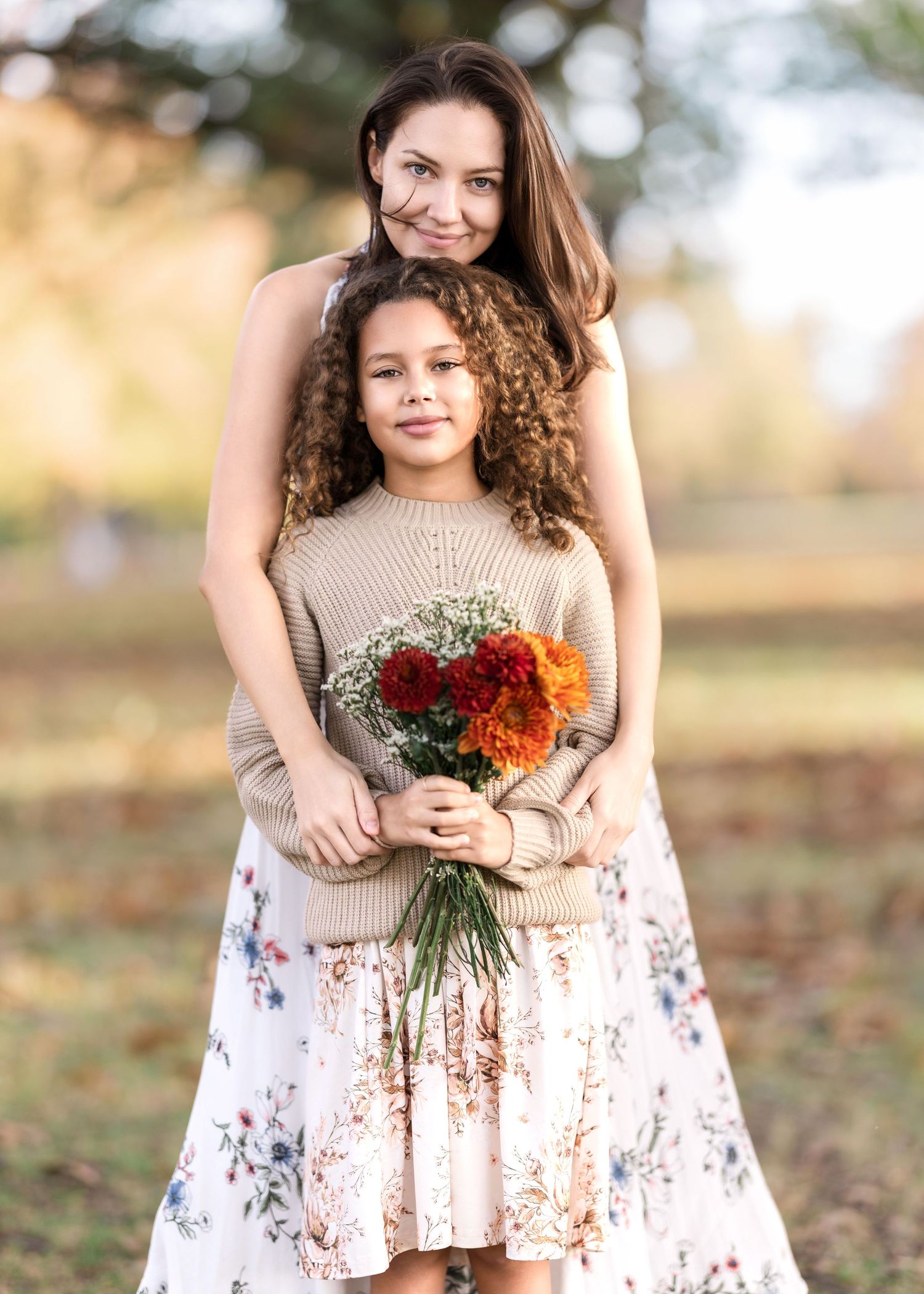 mother embracing daughter and leaning her head on daughter's head from behind.  daughter holding a bouquet of red and orange flowers