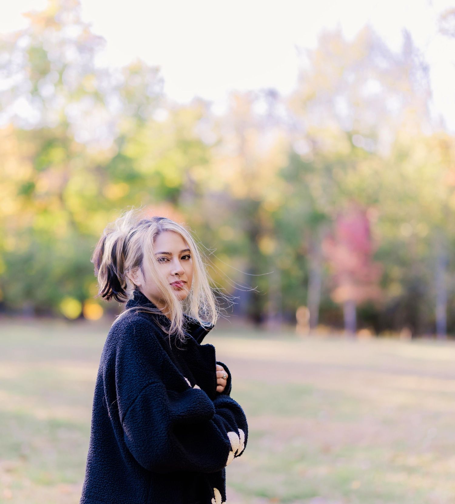 female in the middle of a vast field holding onto her arms while glancing sideways with a dreamy expression
