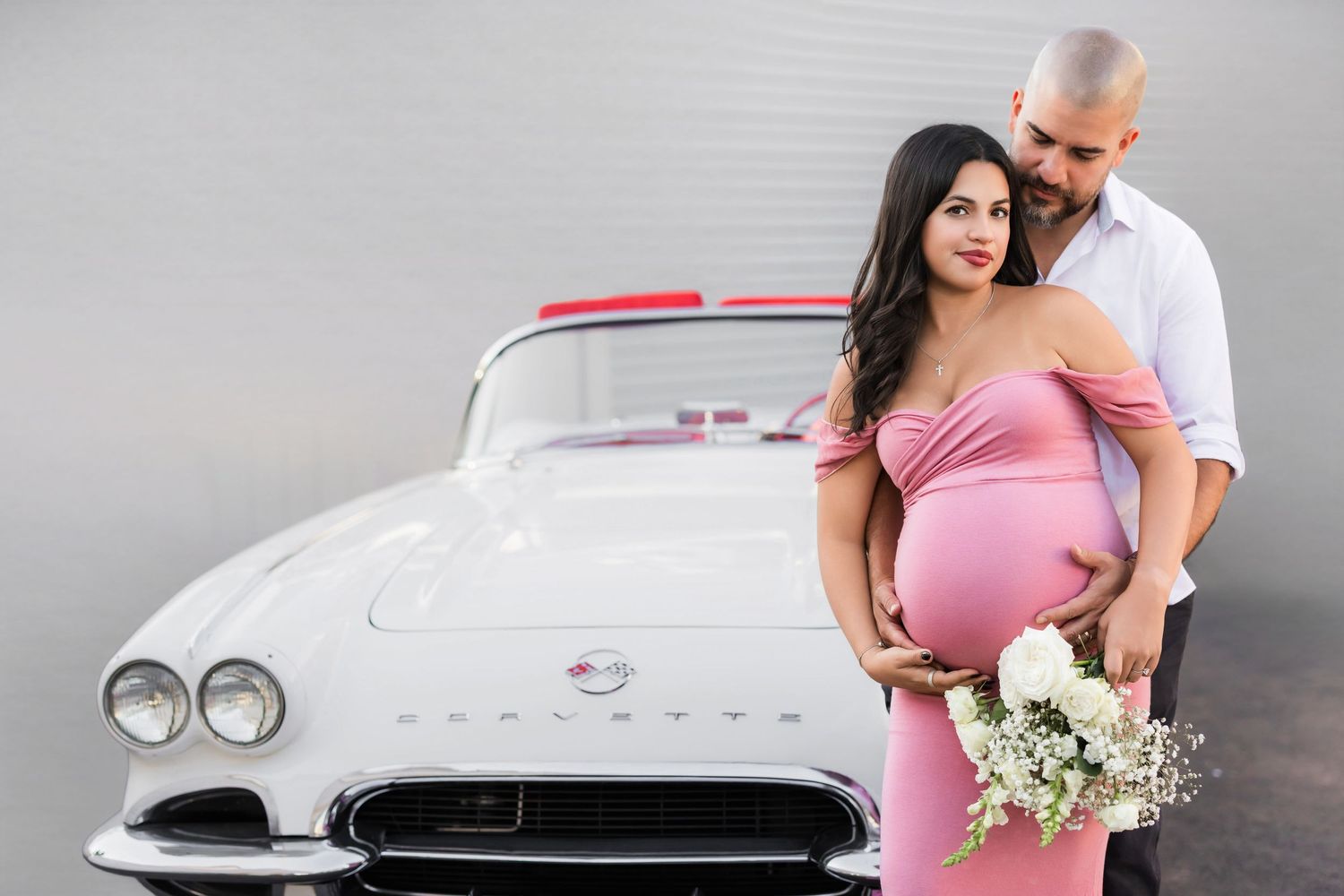female is fitted pink dress and her significant male other stand in front of a class white corvette while holding flowers. maternity shoot