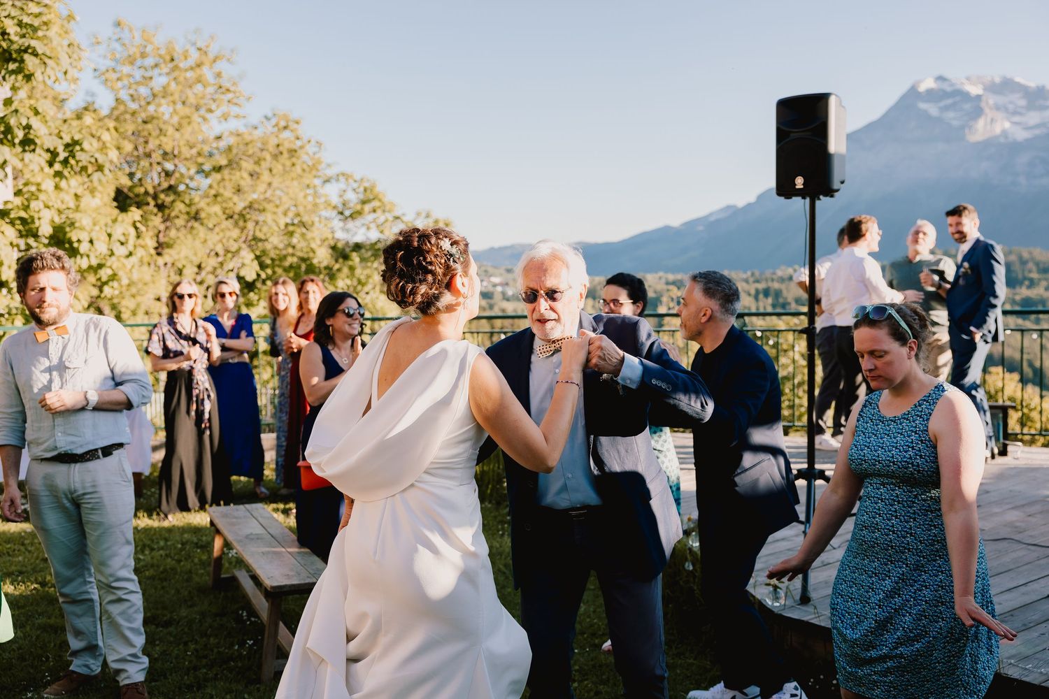 People dancing and celebrating at an outdoor wedding reception with mountain views and a speaker system in the background.