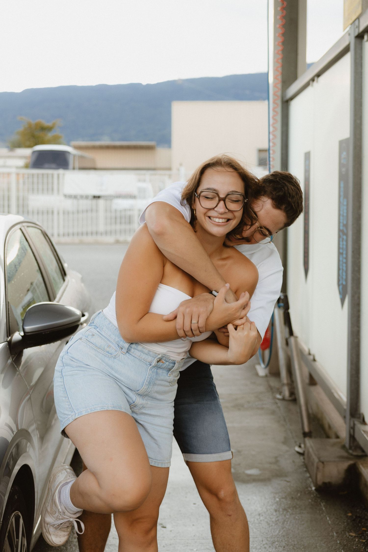 A romantic couple shares playful laughter and embraces in an outdoor parking garage with mountains visible in background.