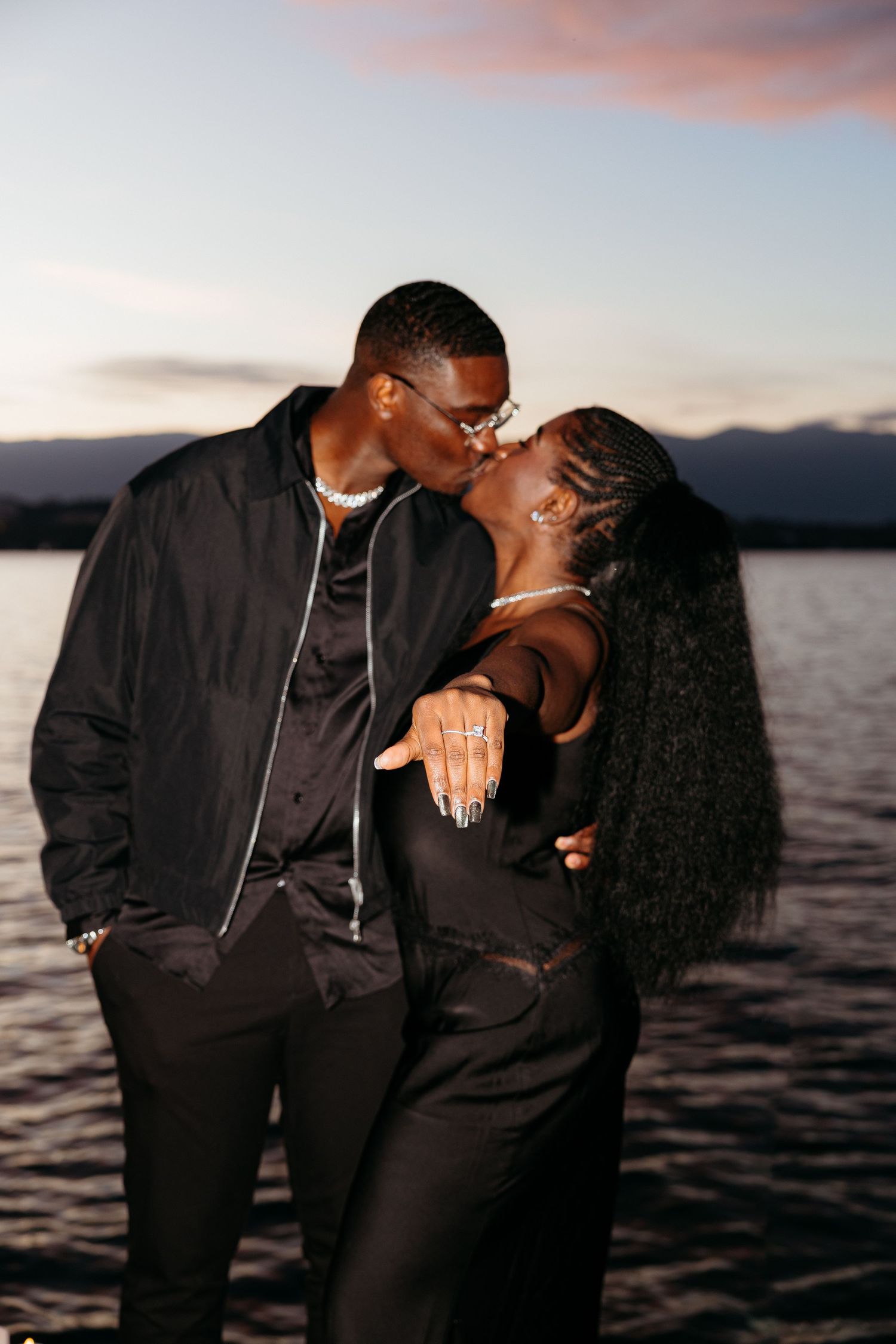 A romantic couple in black outfits share intimate moments by a lake during sunset.