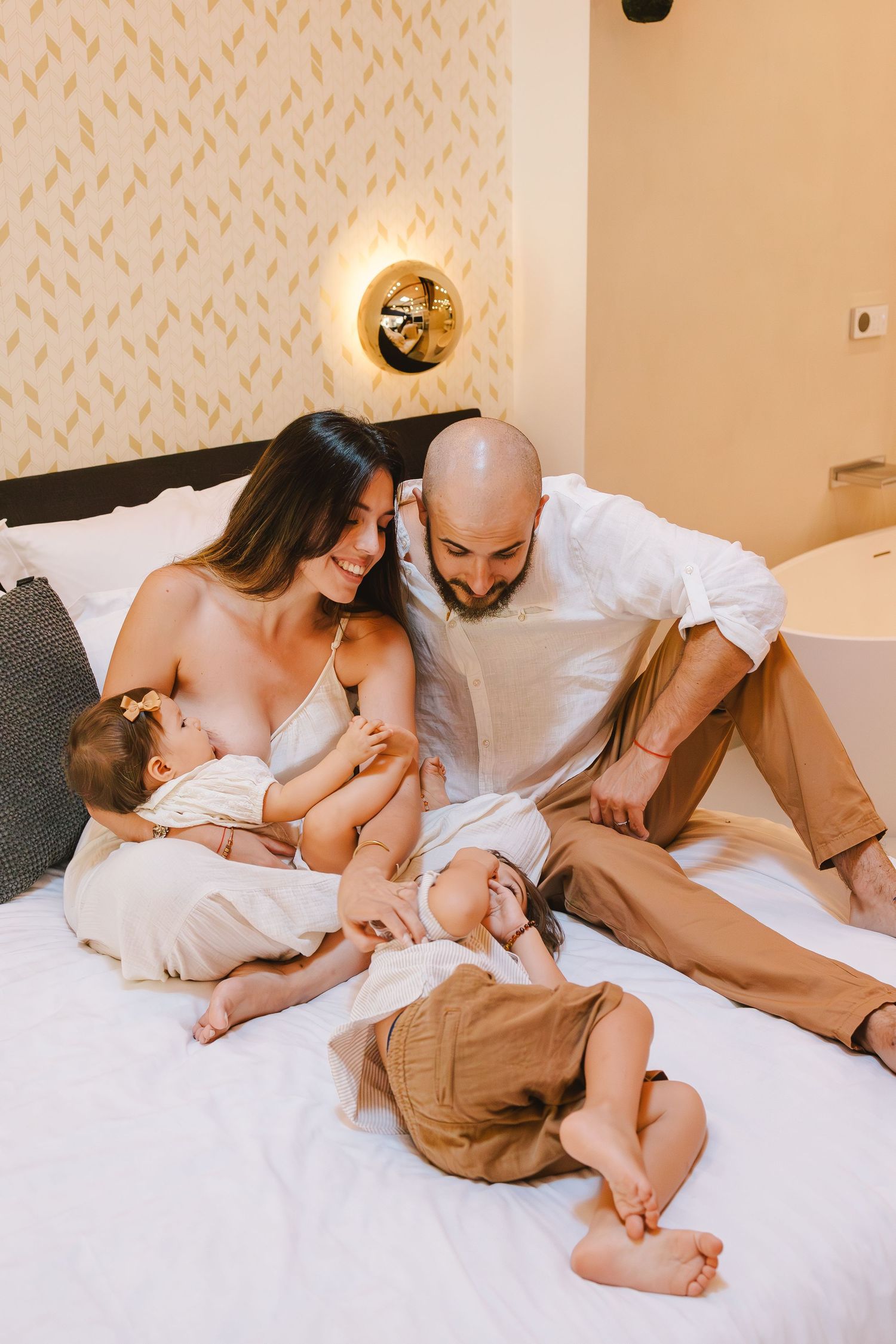 Happy young family with two babies relaxing together on a white bed in a cozy hotel room.