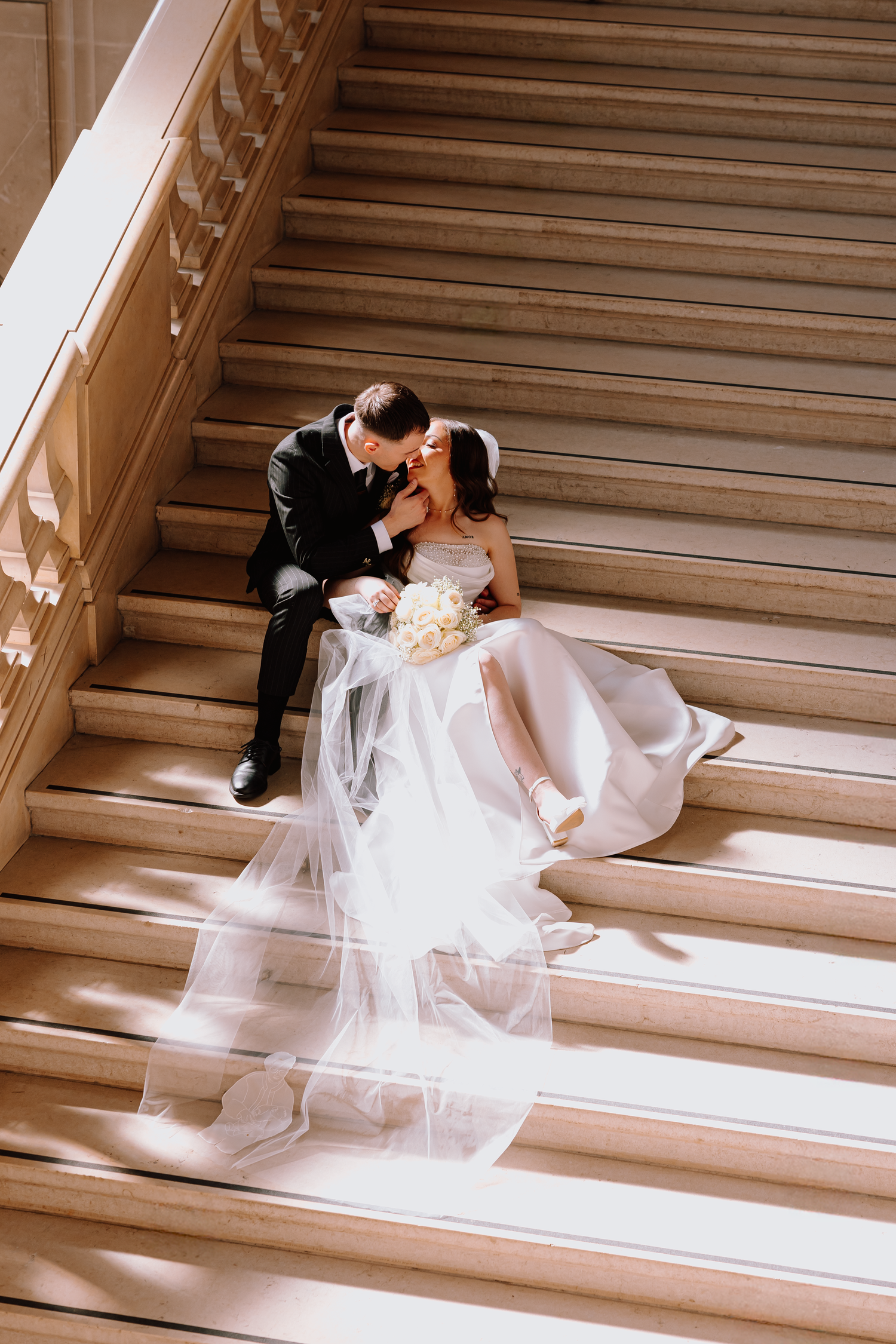 Wedding couple posing dramatically on grand marble stairs in sunlit setting.