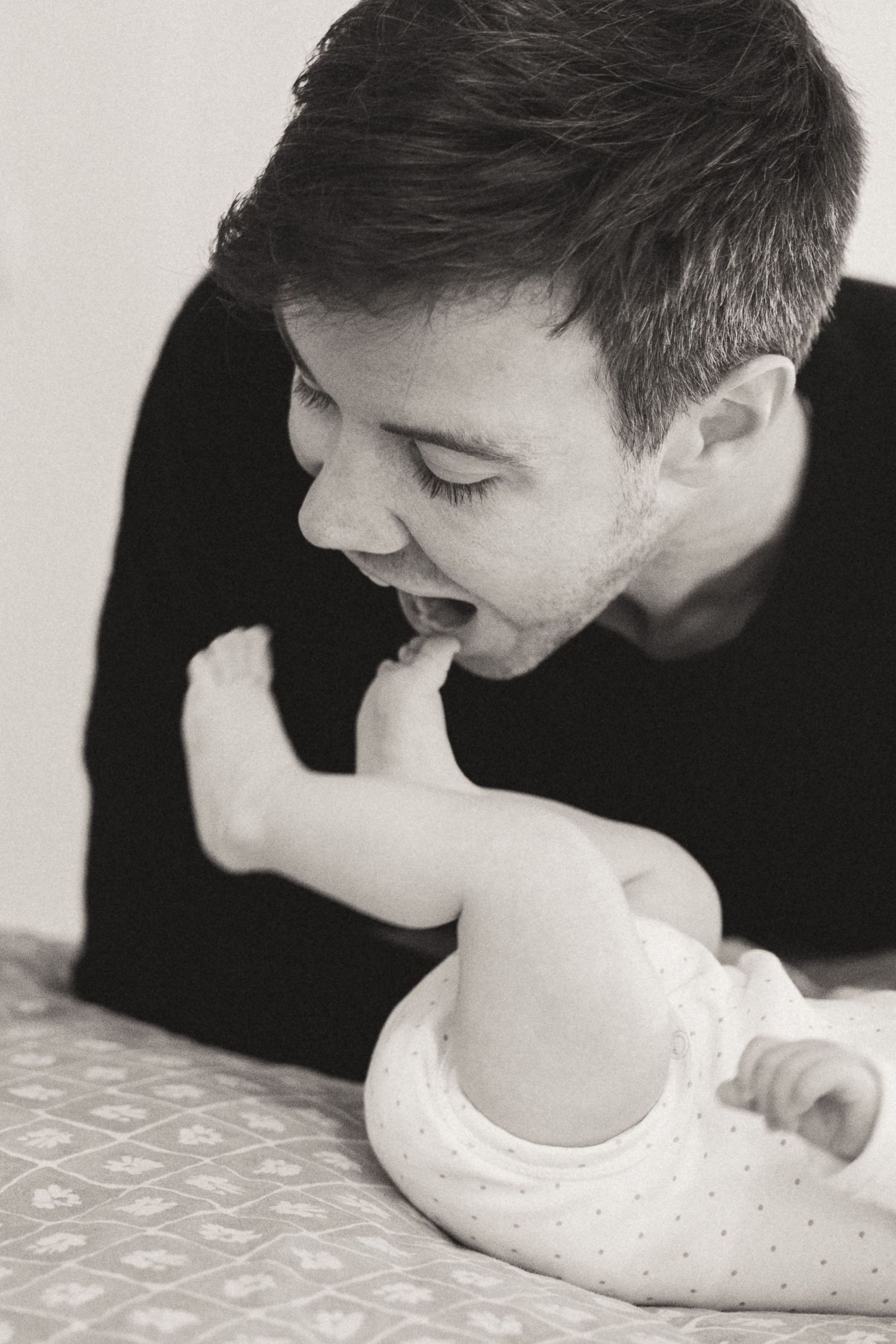 Black and white photo of someone playfully biting a baby's foot while the infant lies on a patterned surface.