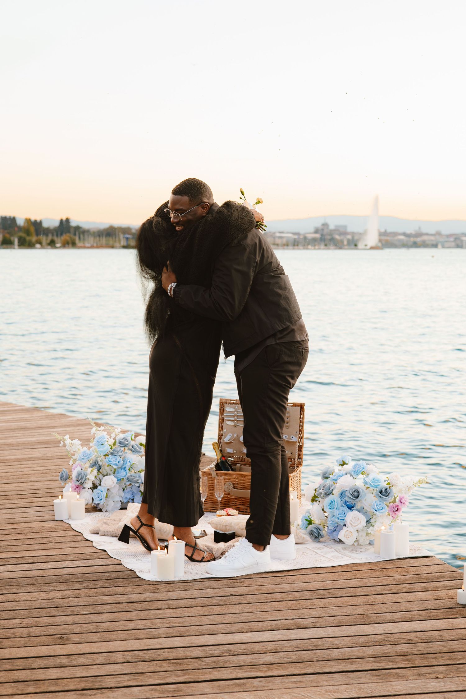 A romantic proposal scene unfolds on a wooden dock by the water during sunset with scattered rose petals.