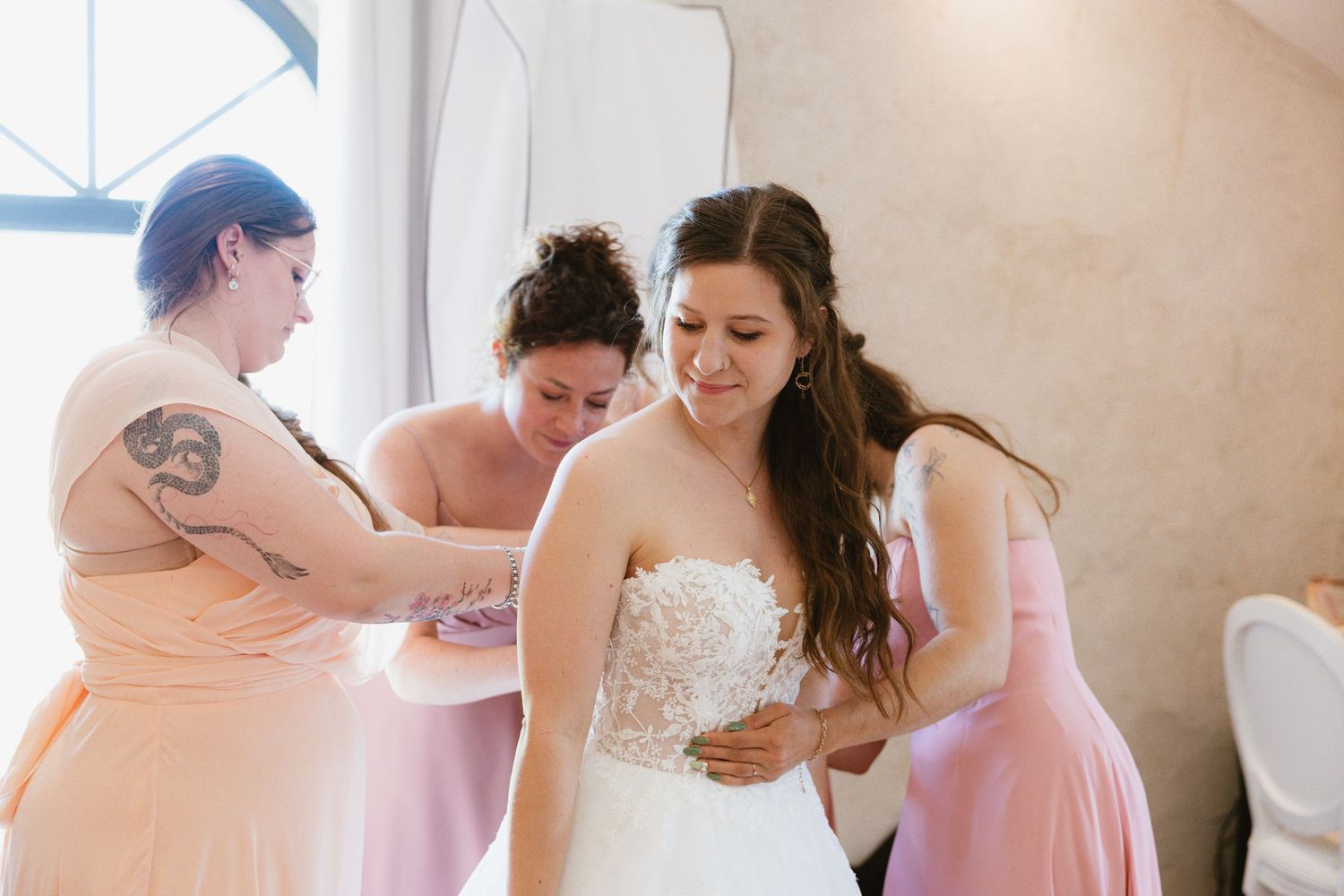 Bridesmaids in pink dresses help the bride get ready in a sunlit room before the wedding ceremony.