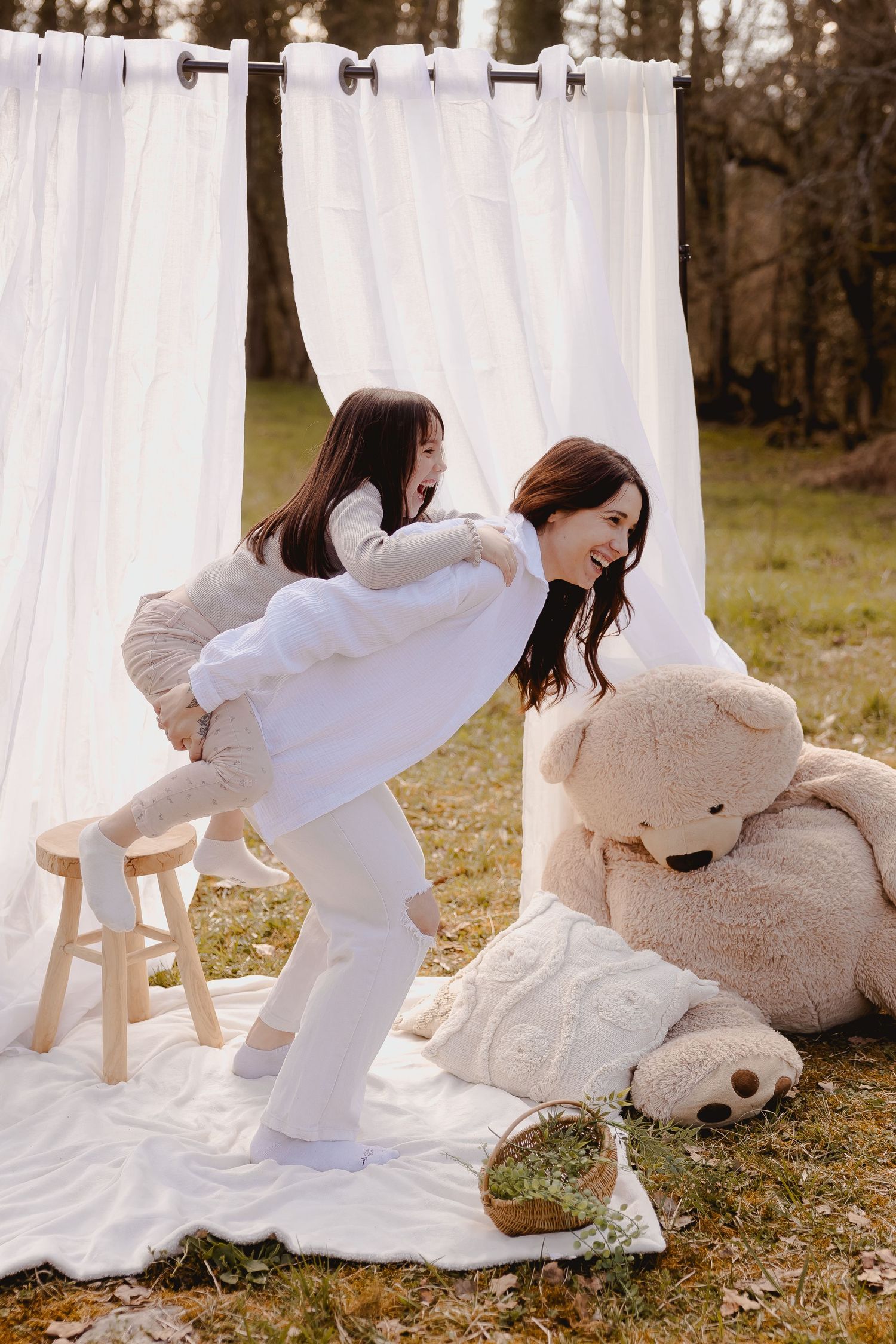 Two people in white outfits playing and laughing outdoors with white curtains and large teddy bear on white blanket.