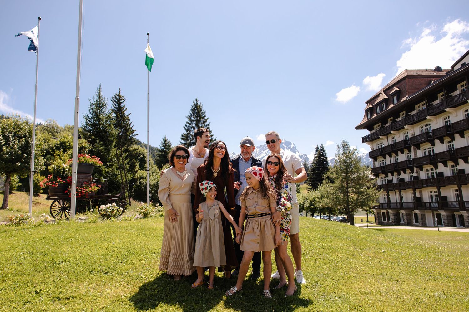 Family group photo outdoors on green lawn near hotel building with flags and pine trees on sunny day.