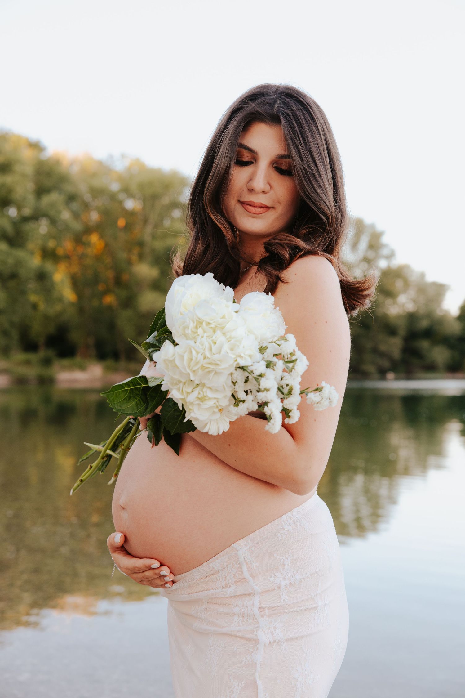 A set of maternity photos show a woman in a blush dress holding white flowers by a serene lakeside at sunset.