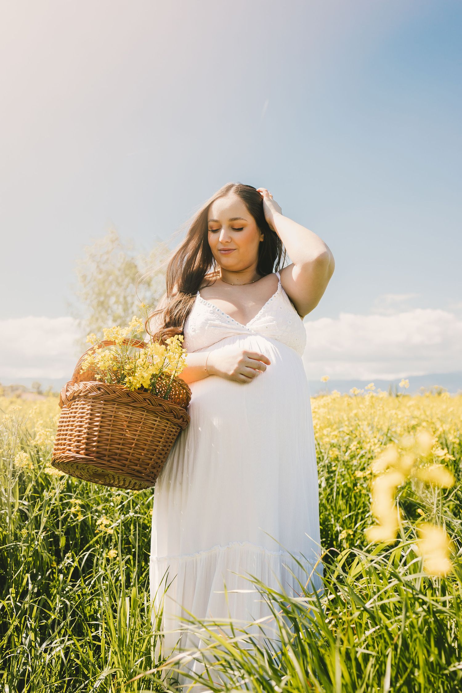Pregnant woman in white dress holding flower basket in sunny yellow field.