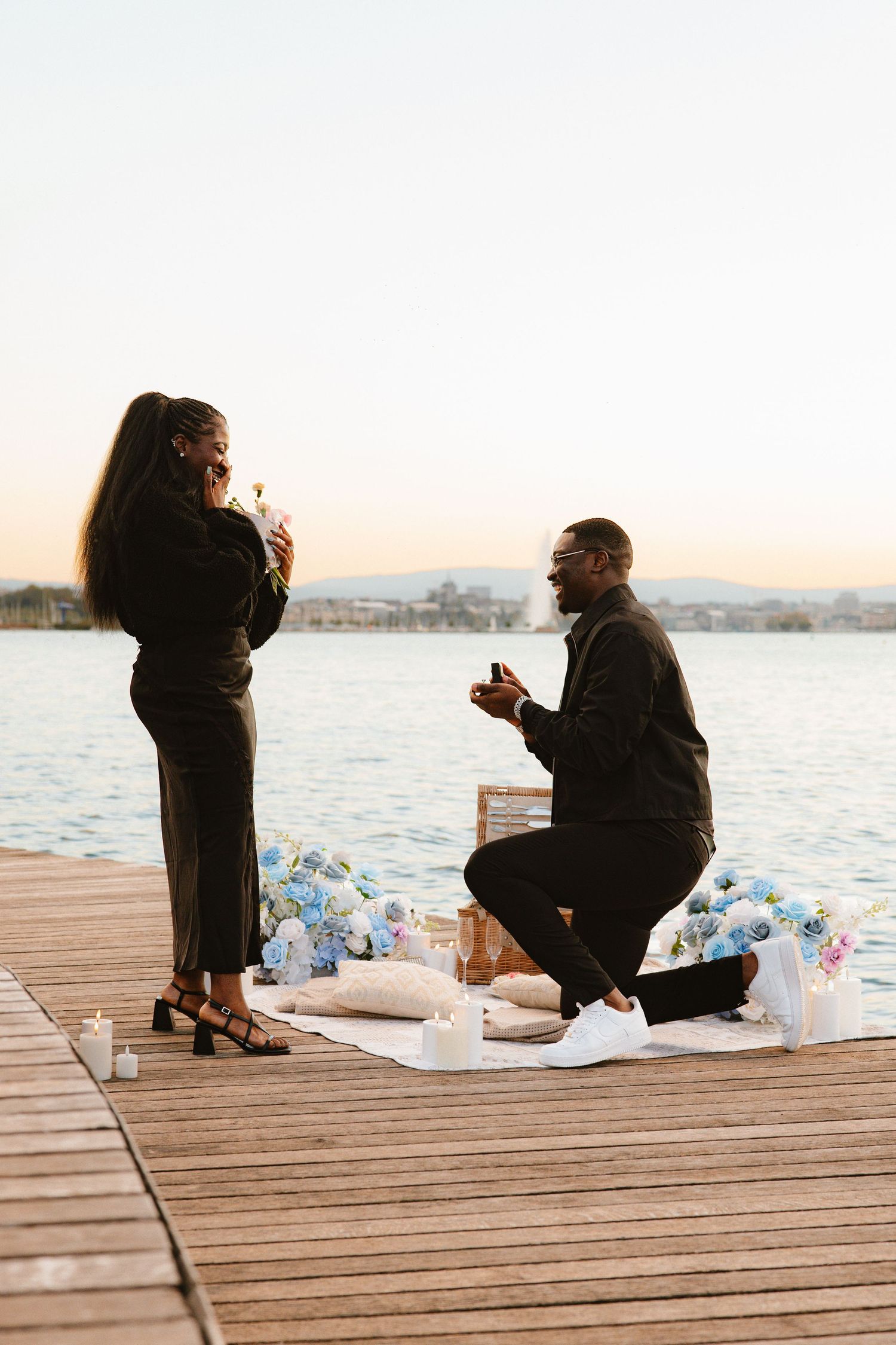 Two people sit together on a wooden dock by the water at sunset sharing a romantic moment.