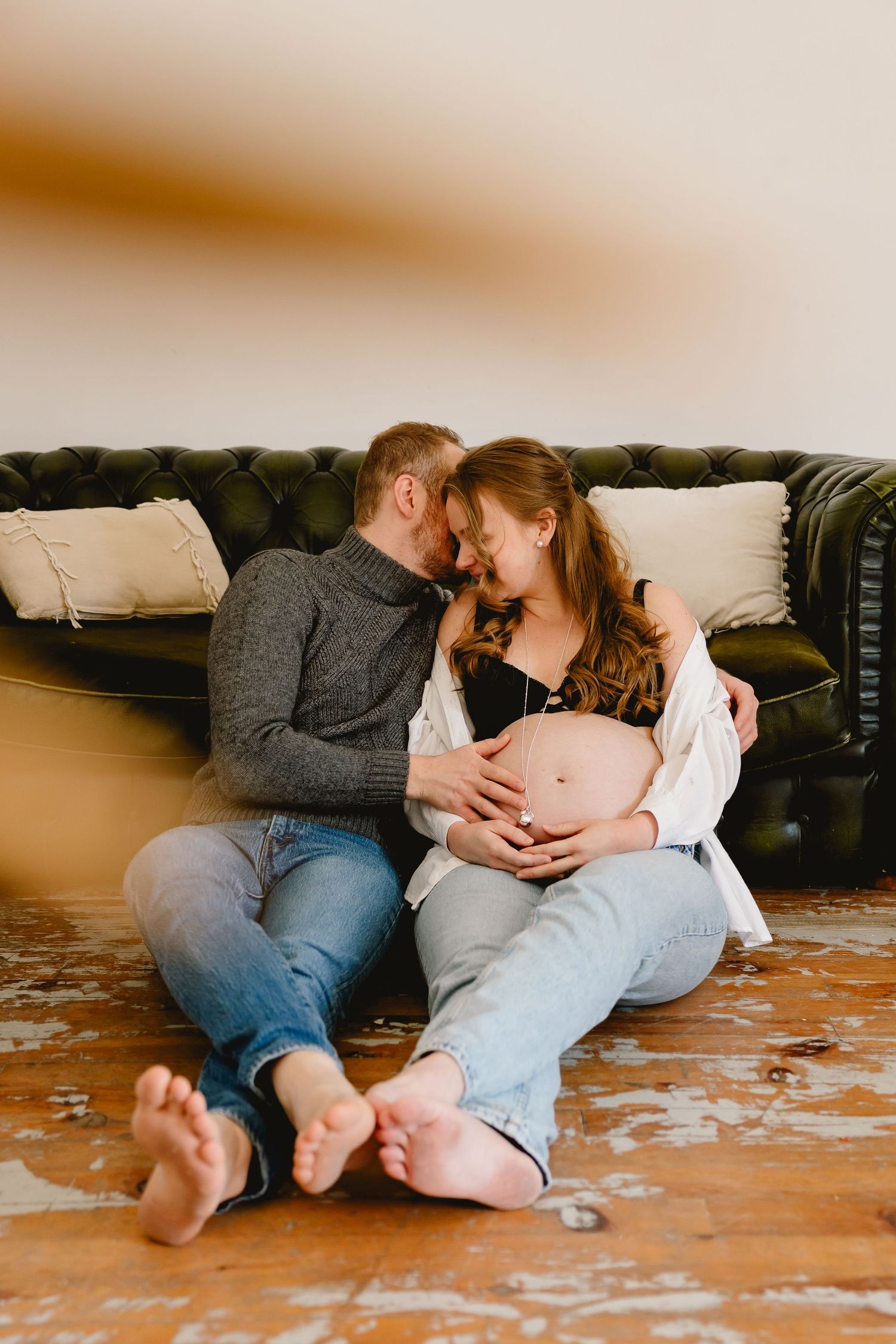 Expecting couple sits intimately on floor against dark leather couch, hands resting on pregnant belly.