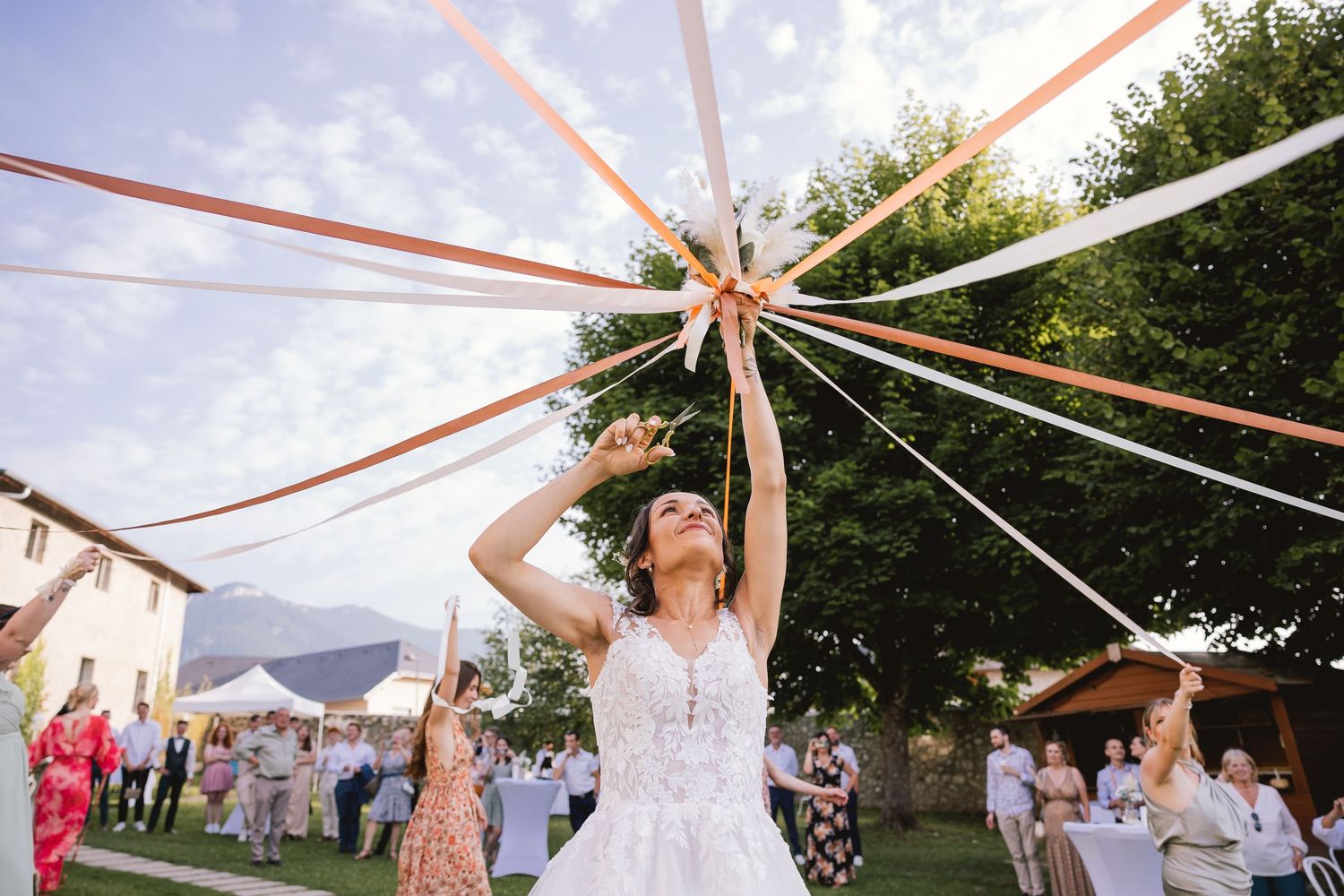 A festive outdoor wedding celebration with colorful ribbons held up in a maypole style under a bright blue sky.