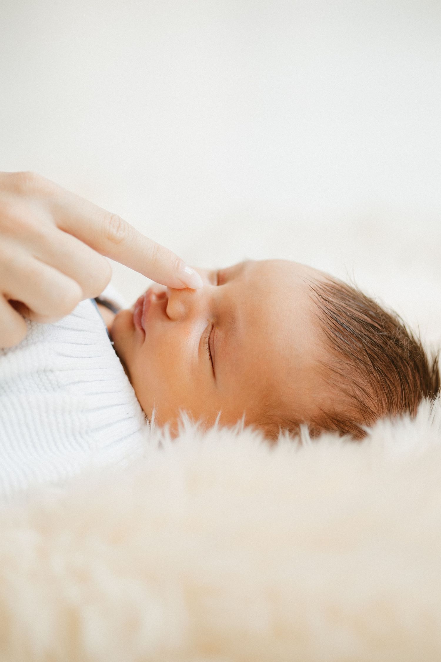 A sleeping baby wearing white lies peacefully on soft white fur bedding.