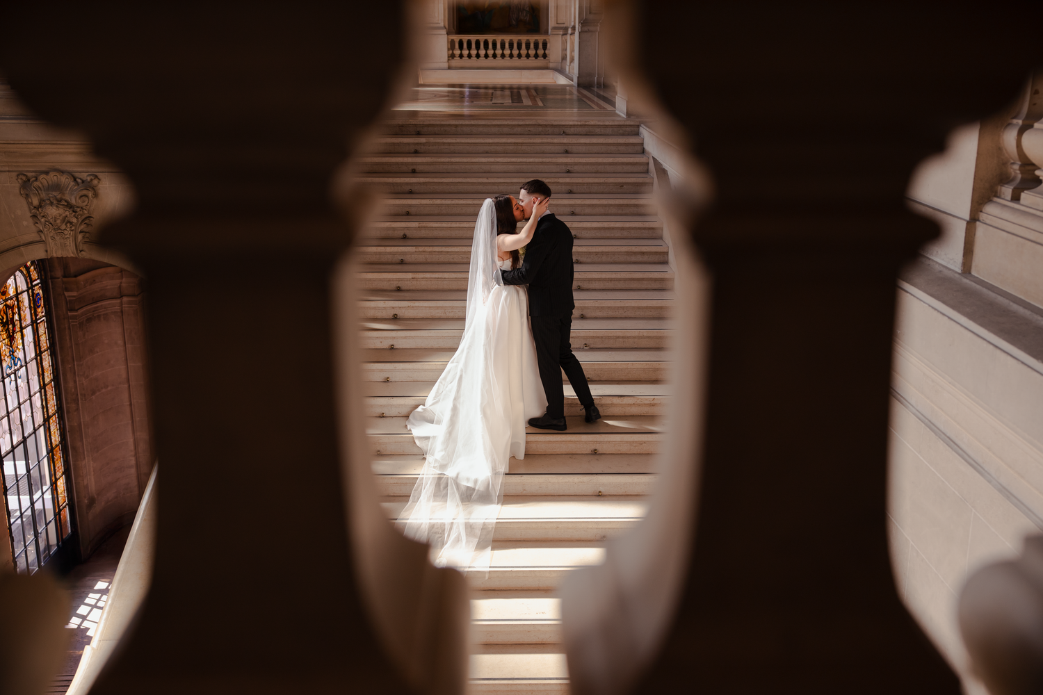 Bride and groom share a romantic kiss on grand staircase, framed by elegant stone archway in ornate wedding venue.
