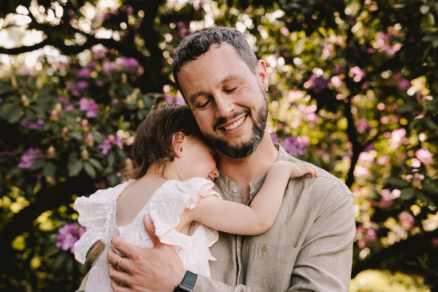A smiling bearded father lovingly embraces his baby girl outdoors near blooming purple flowers.