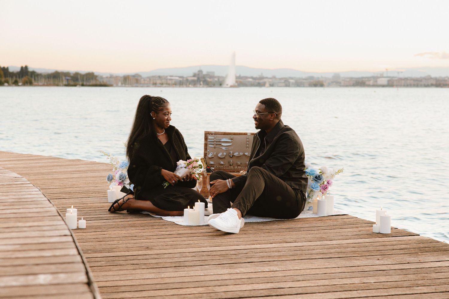 Two people sit together on a wooden dock overlooking a serene lake during sunset.