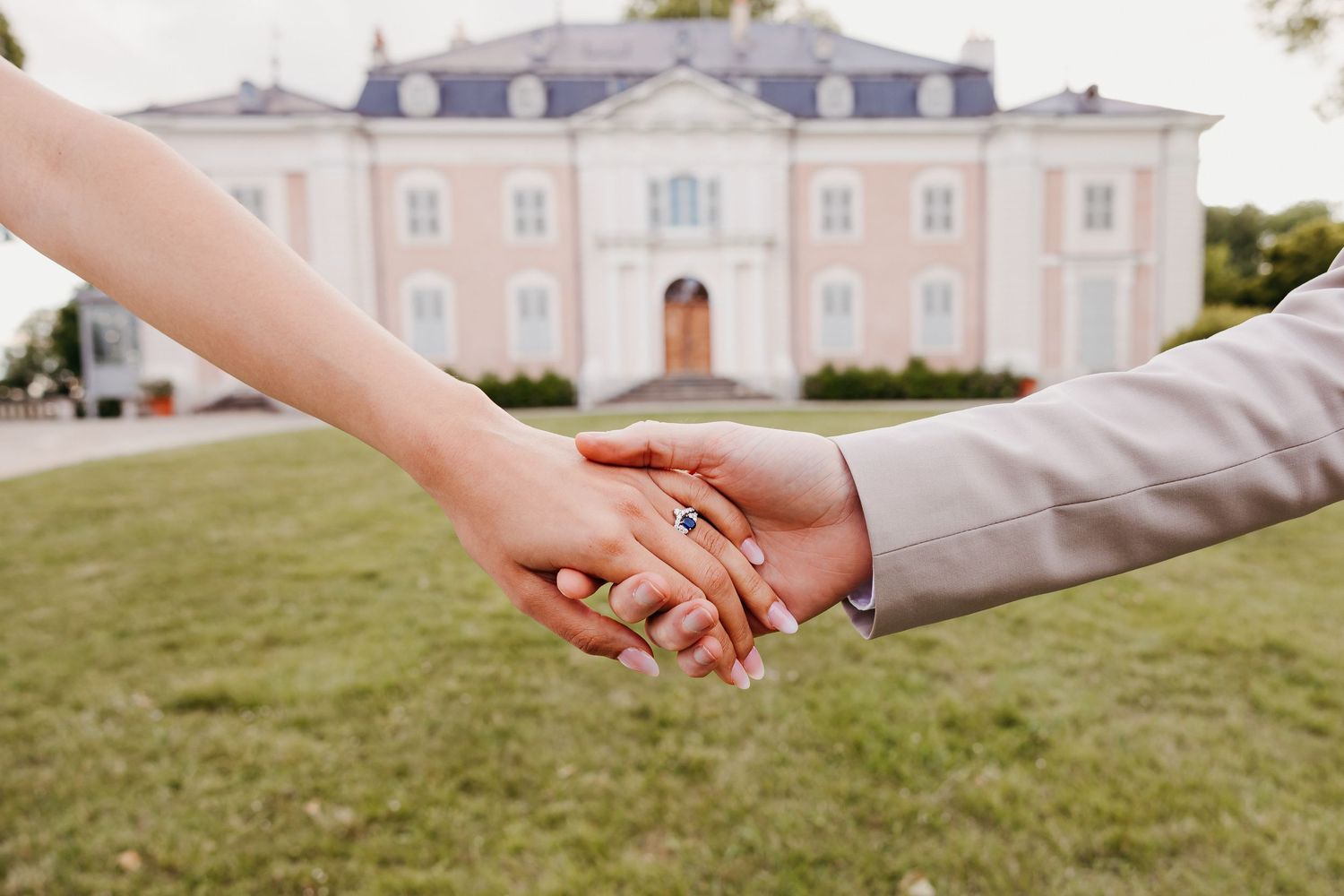 Two hands clasped together in front of an elegant pink mansion with a manicured lawn.