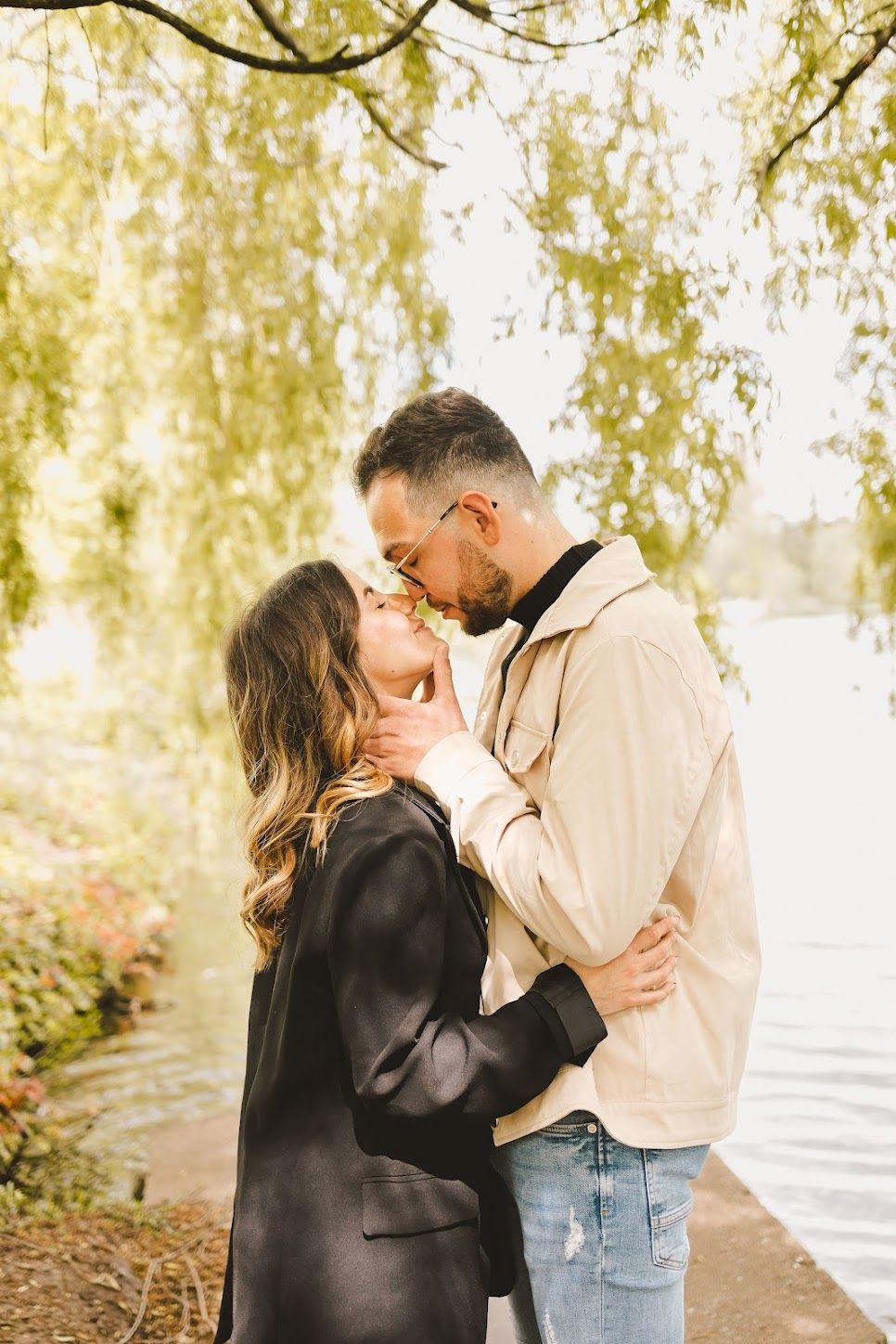 Couple sharing a romantic kiss near a serene lake under a weeping willow tree in autumn.