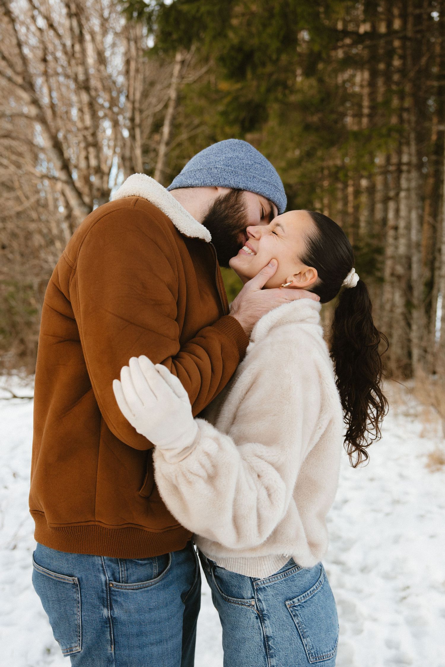A couple shares a romantic moment in a snowy winter forest while wearing casual clothing and warm sweaters.