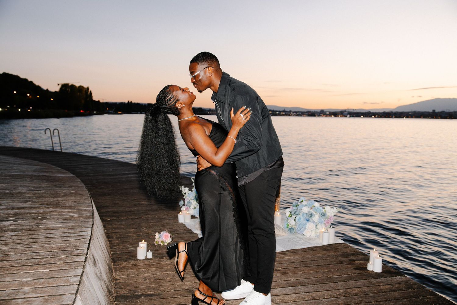 A romantic couple embraces on a wooden dock at sunset with candles and a serene lake view in the background.