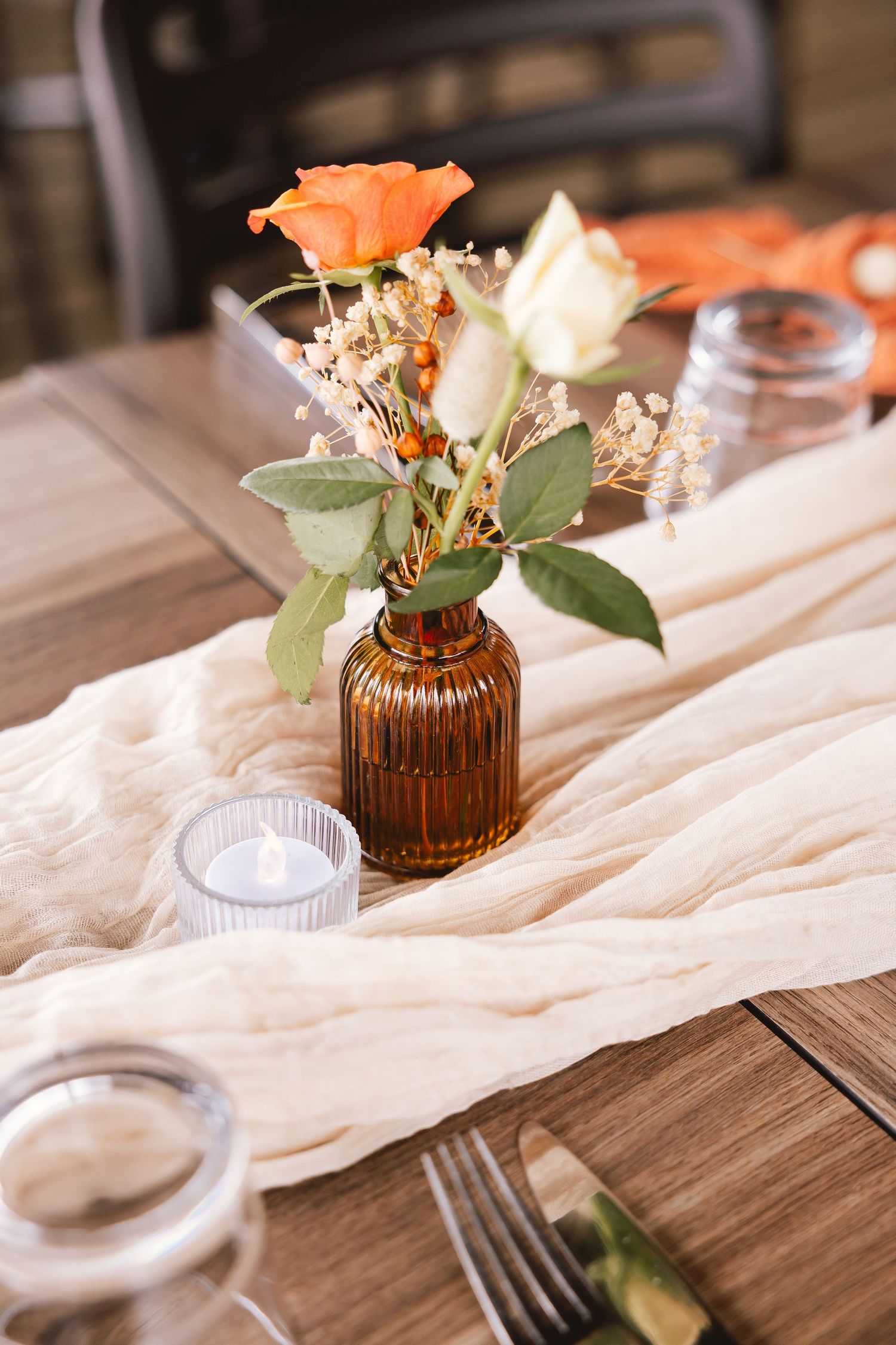 A small amber vase holds orange roses and white flowers on a wooden table with gauzy fabric and candle holders.