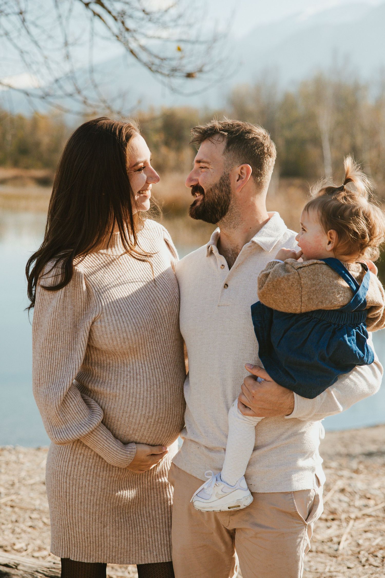 Family wearing neutral tones embraces and laughs together during outdoor photoshoot by a lake on autumn day.