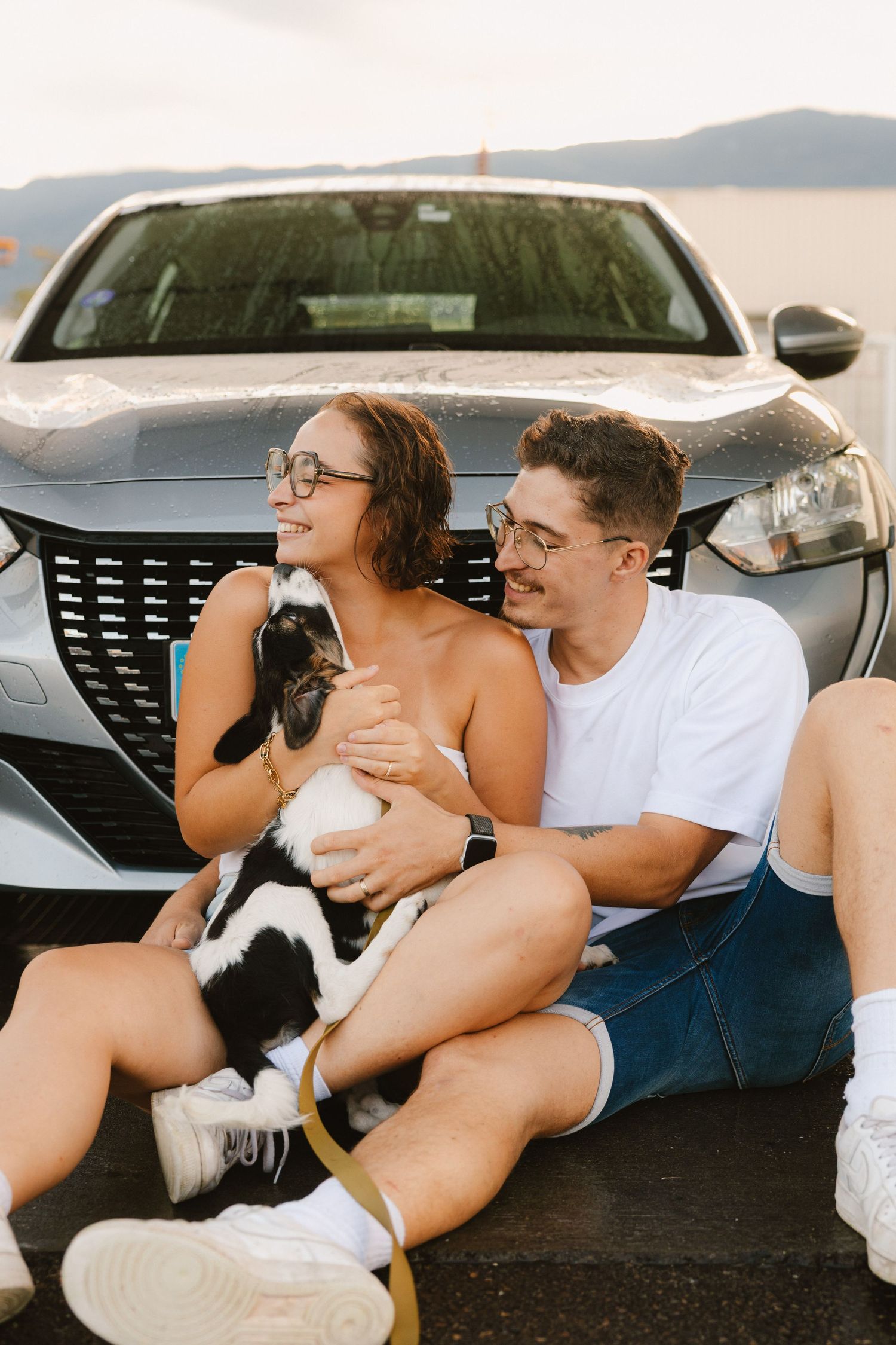 A couple sits together in front of a silver car while holding a black and white pet dog.