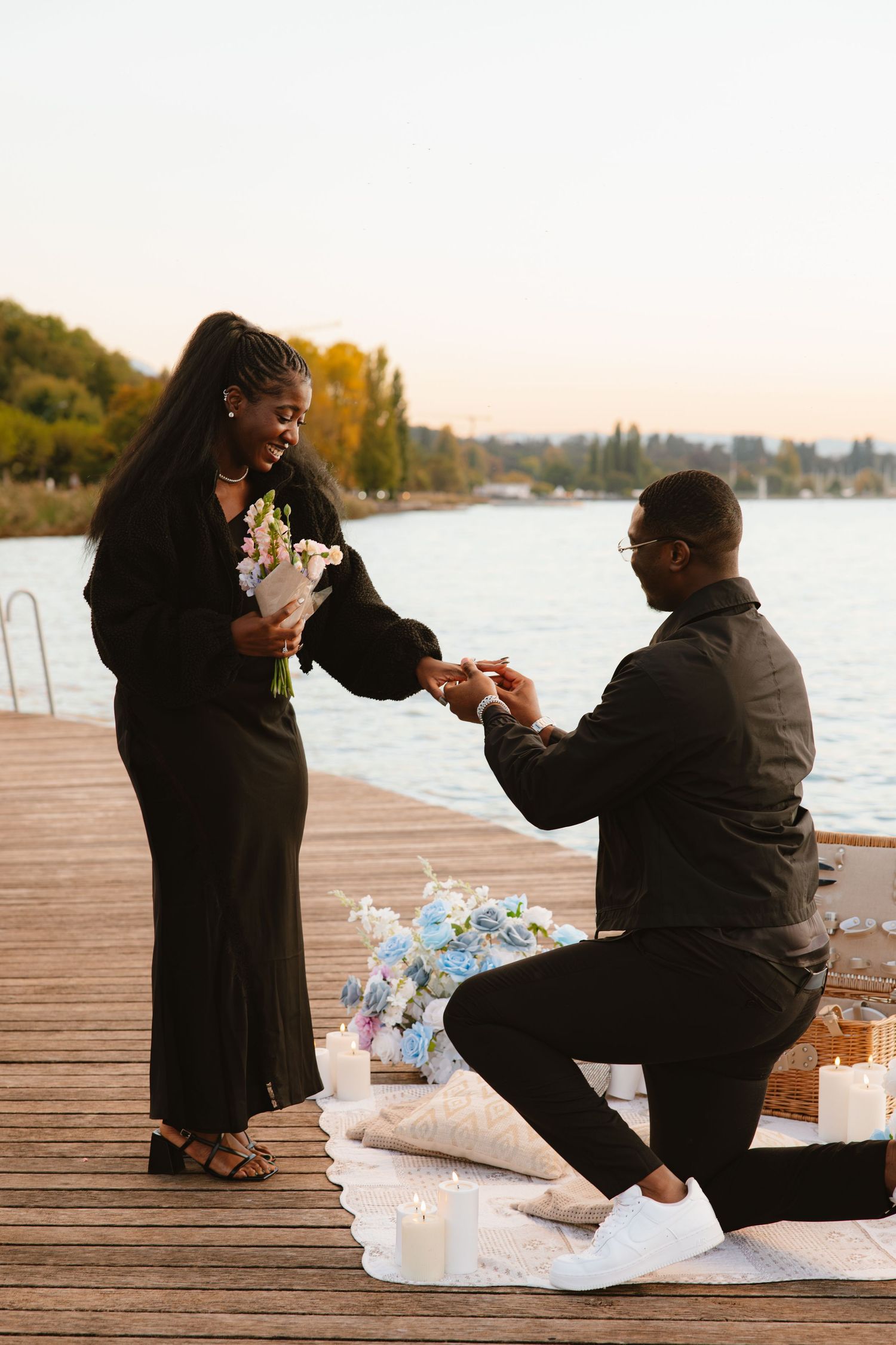 A romantic proposal sequence on a lakeside dock at sunset with decorative lights and flowers.