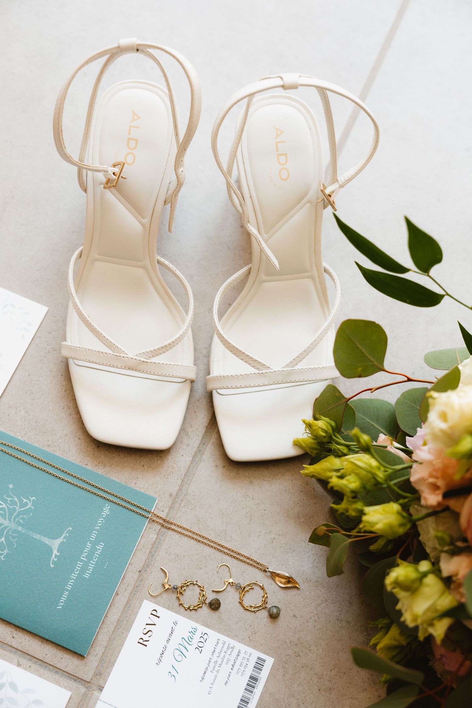 Elegant white bridal sandals with delicate straps displayed next to a teal book, jewelry, and floral arrangement.