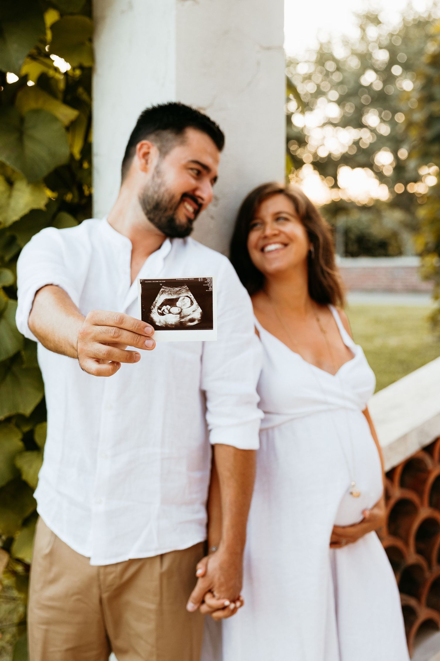 A loving maternity photoshoot outdoors shows a couple in white outfits embracing against a dreamy sunlit background.