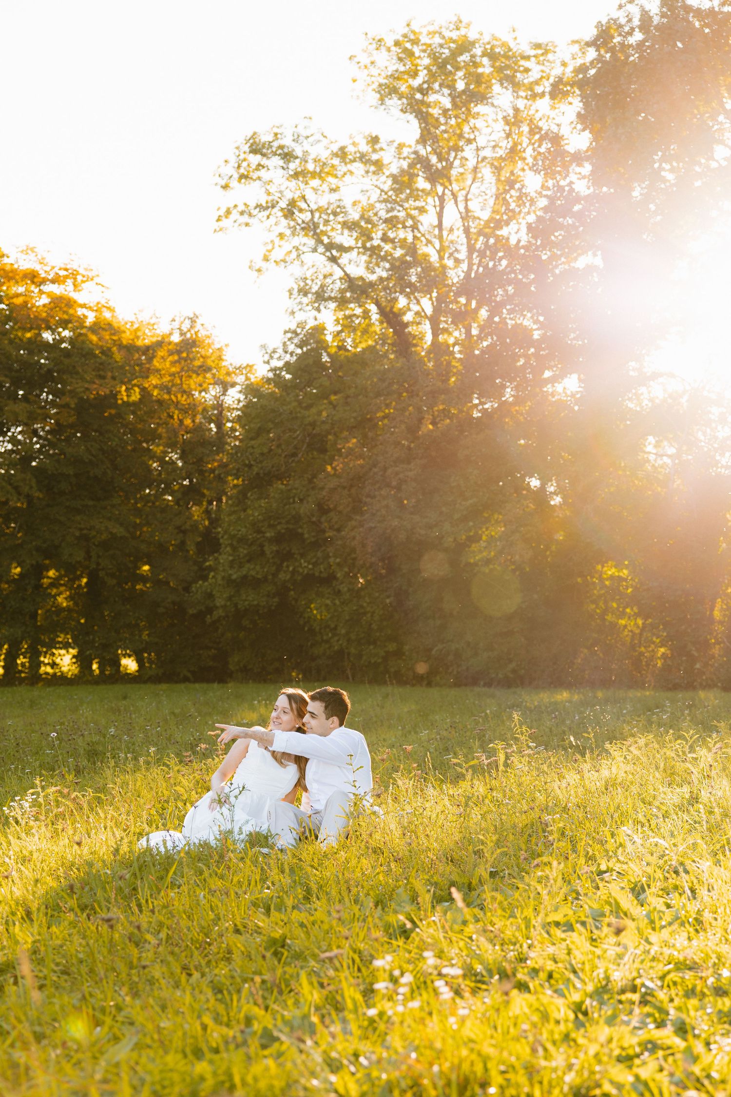 Two people in white clothing sit together on a grassy hill during golden hour sunset with trees in the background.