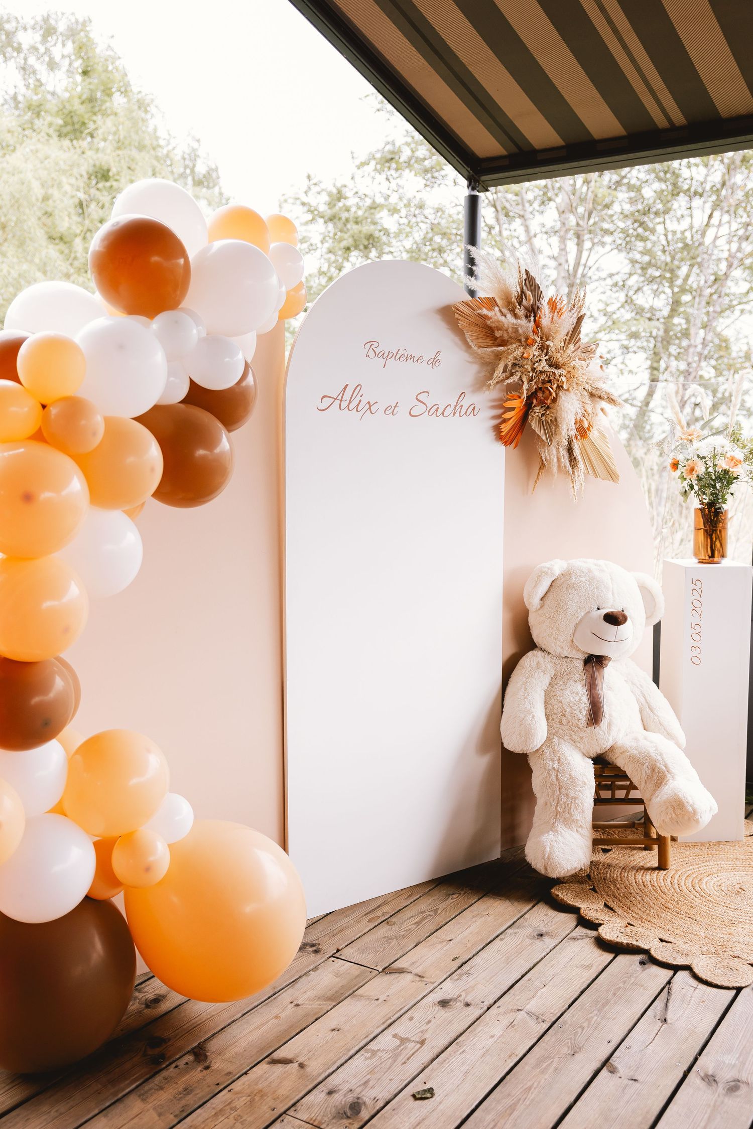 A rustic patio decorated with orange and white balloons featuring a large teddy bear against a white backdrop for celebrations.