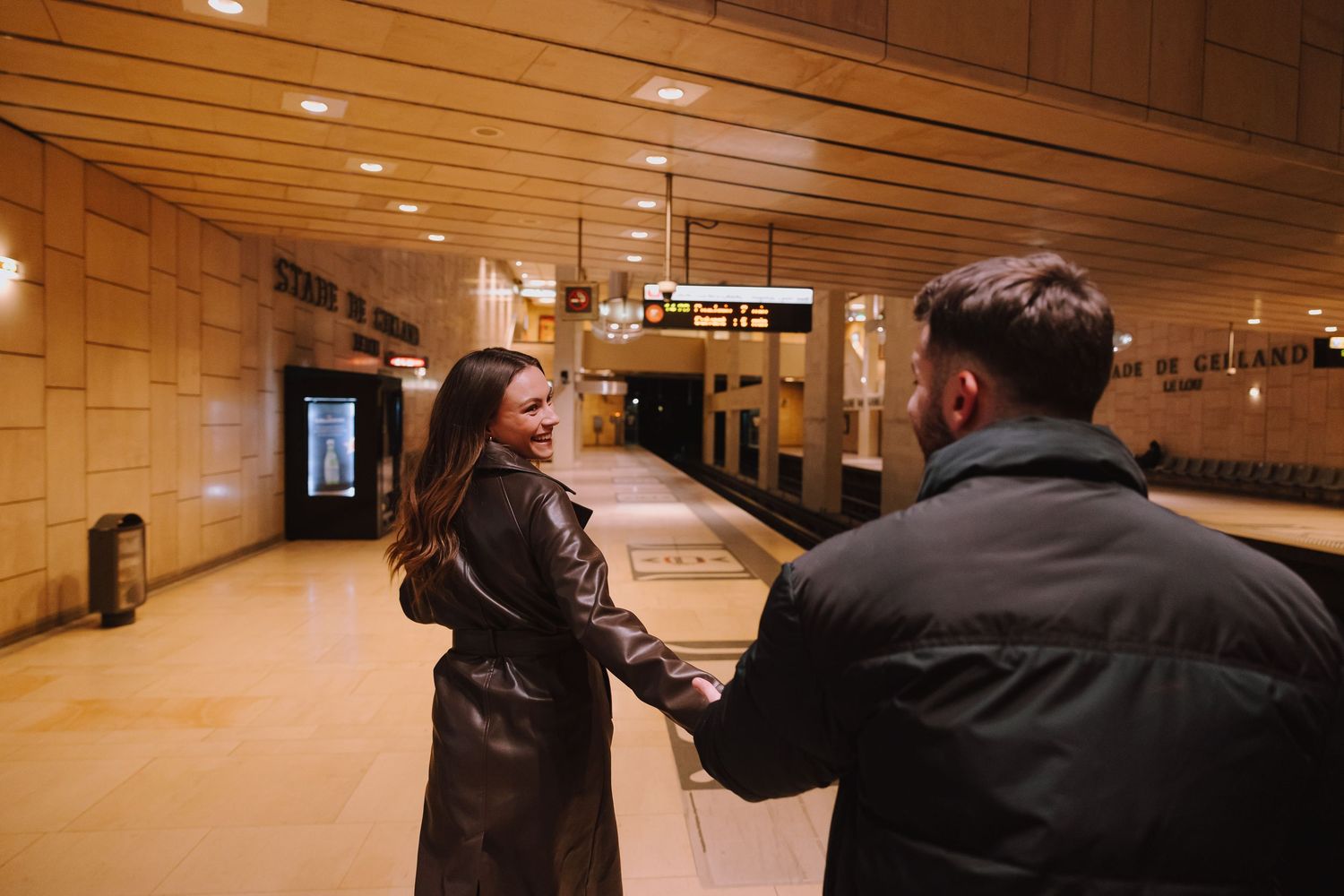 Two people standing in a warmly-lit empty subway station with wooden panels on the ceiling.