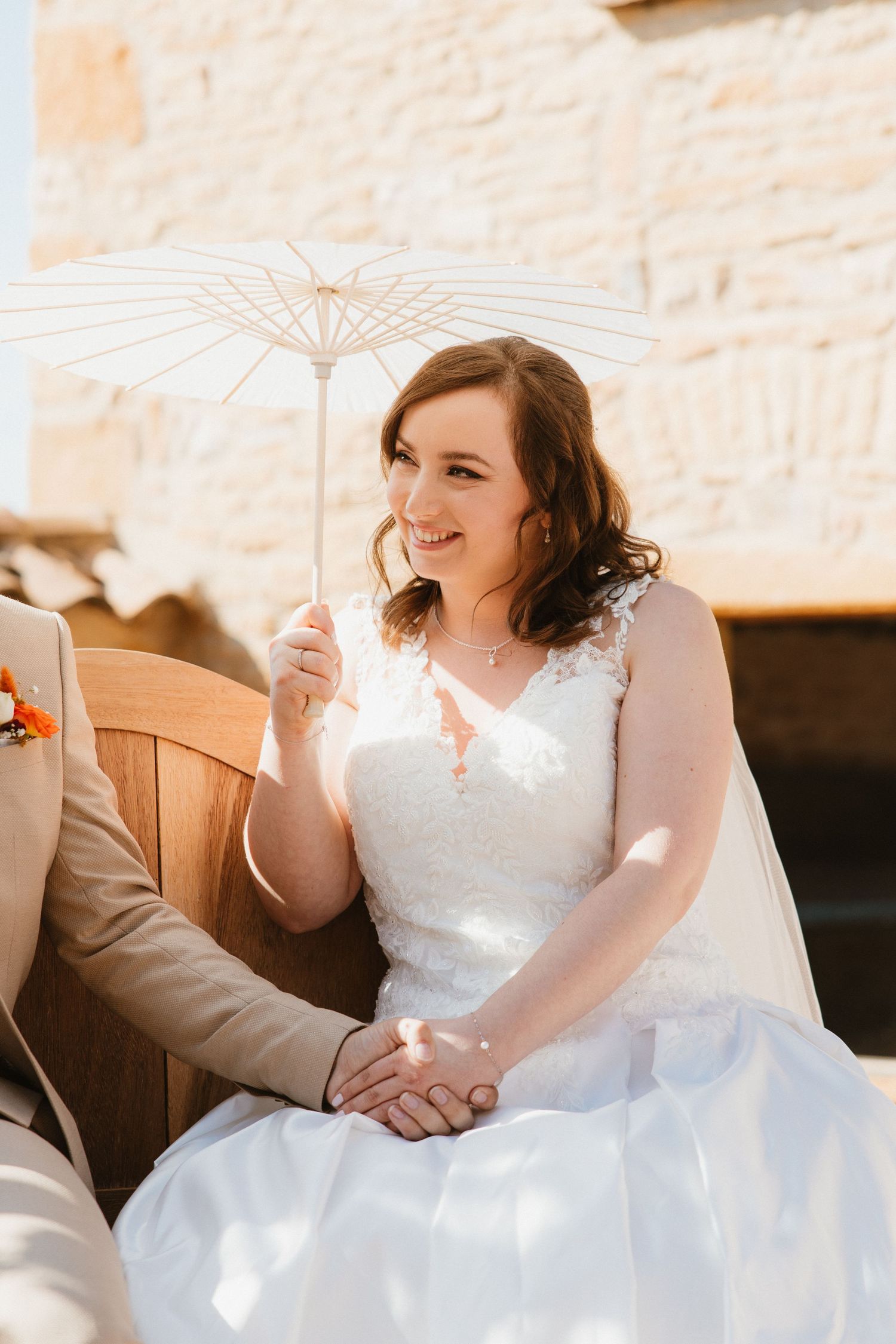 A person in a white wedding dress sits on a wooden bench holding a delicate white parasol in bright sunlight.
