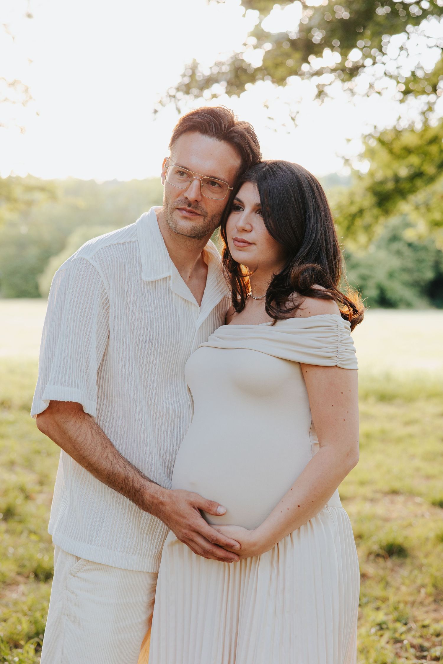A couple in white outfits embrace during a maternity photoshoot in a sunlit outdoor setting at golden hour.
