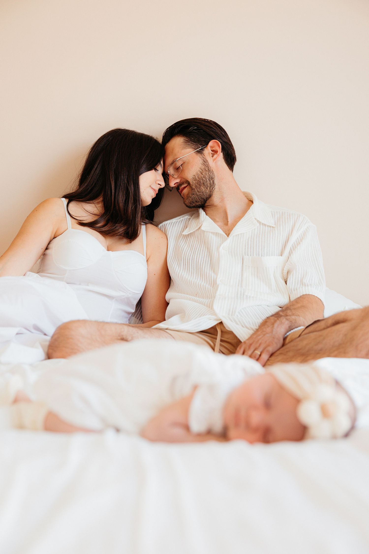 A couple rests together in white bedding showing intimacy and connection in a bright bedroom setting.