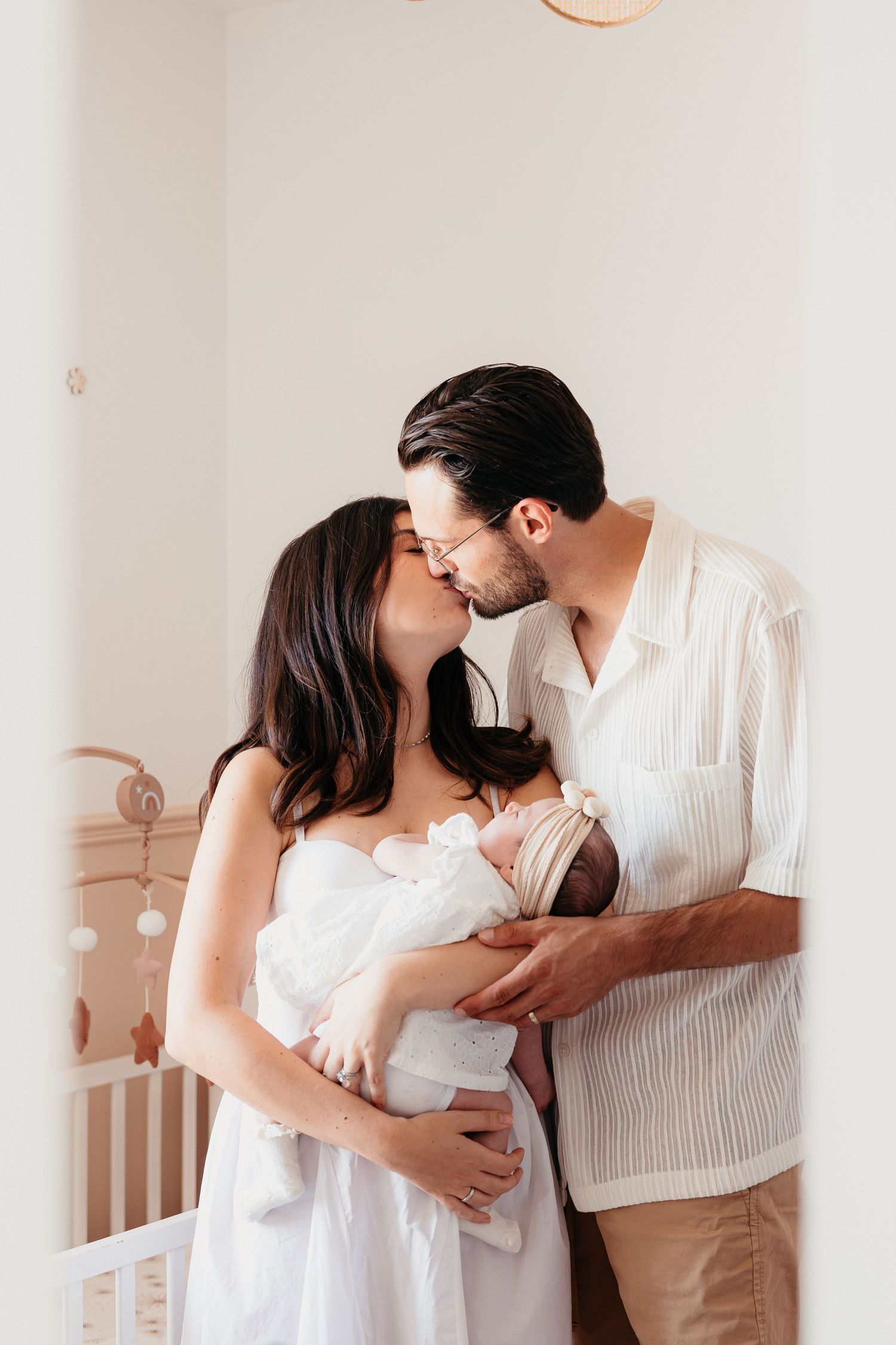 A couple in white clothing share an intimate moment while holding their newborn baby in a nursery setting.
